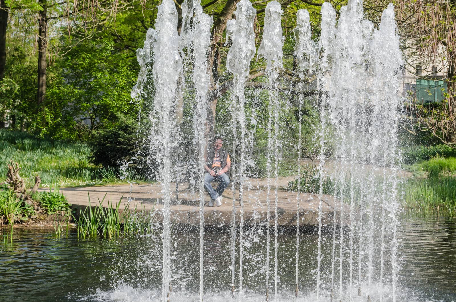 Water wall behind the man sitting on a bench, created by a fountain fountain in a public park.
