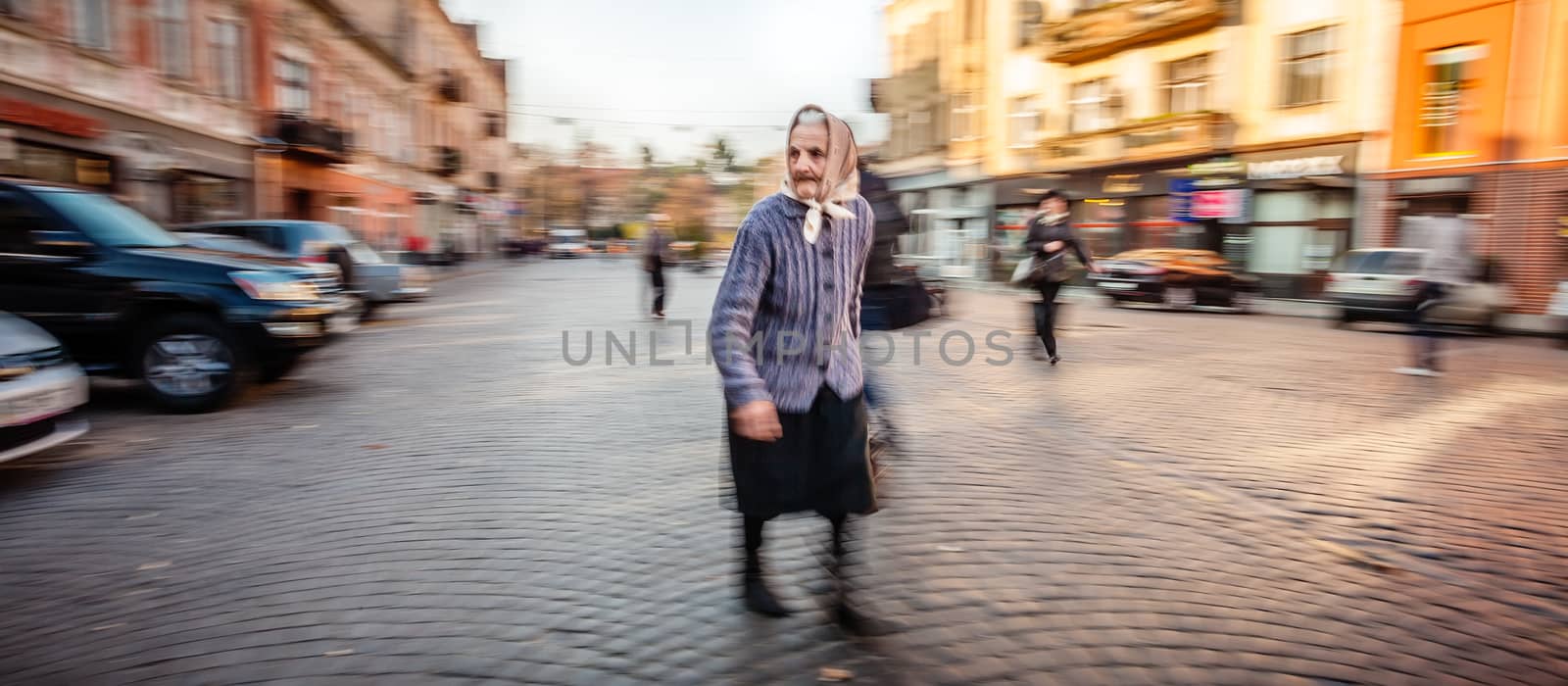 UZHGOROD, UKRAINE - OCT 25, 2013:  lonely senior woman in a shawl and wool sweater walking in a paved city square in the early morning. Intentional motion blur