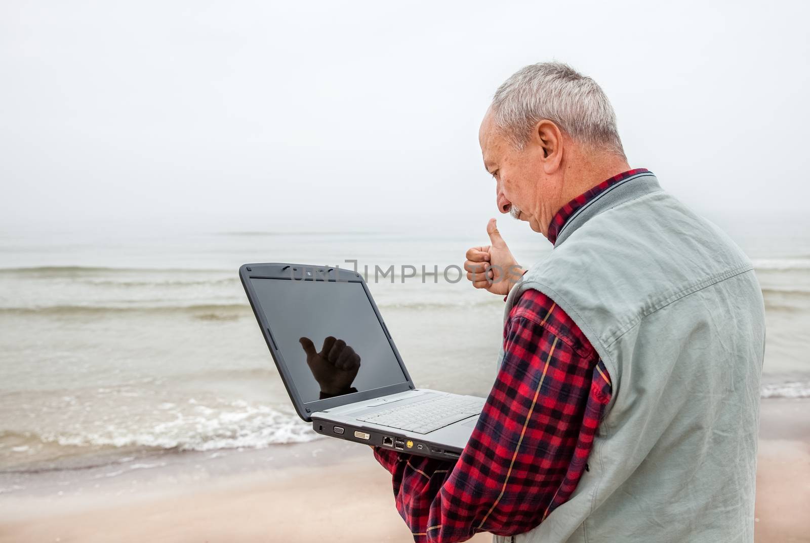 All OK. Old man standing on the beach with a laptop and a raised thumb up.