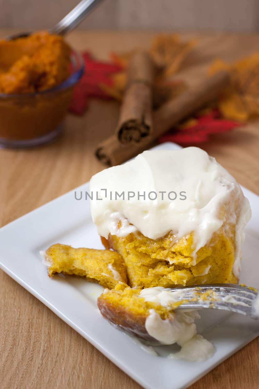 Homemade pumpkin cinnamon roll on a white plate with pumpkin, cinnamon, and fall leaves on a wooden table.