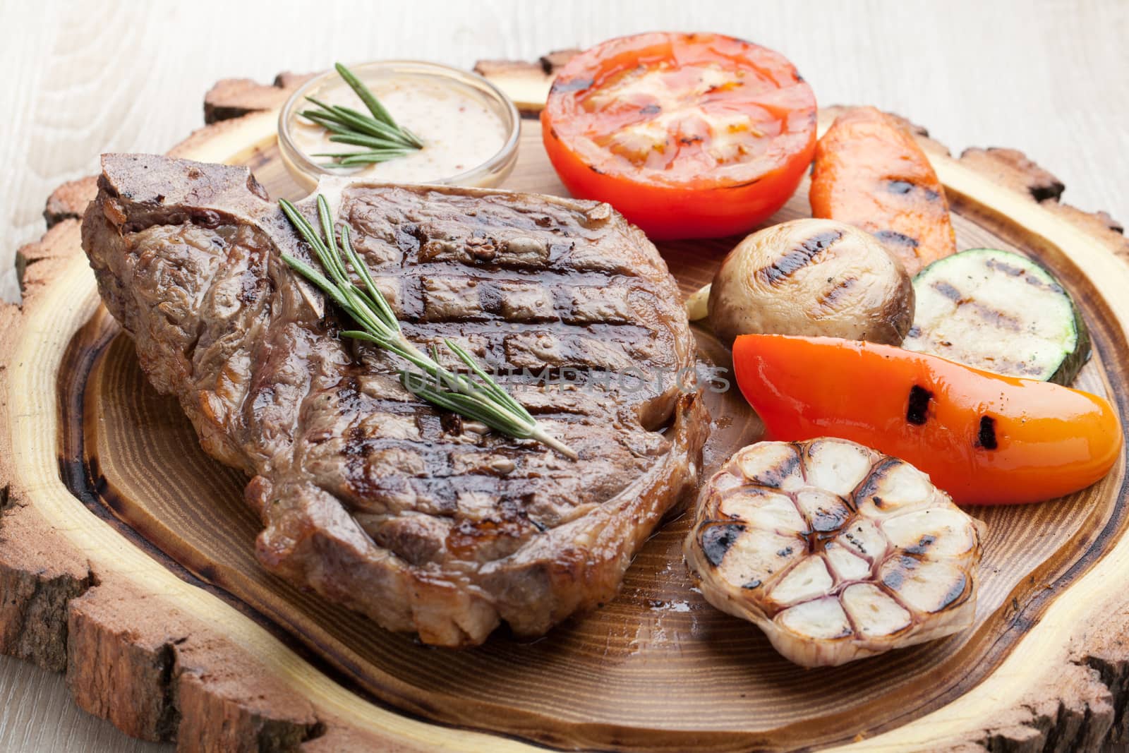 Portion of BBQ t-bone steak  served  on wooden board with  rosemary, mustard sauce  and grilled vegetables : tomato, carrot, paprika, garlic,  champignon,  zucchini