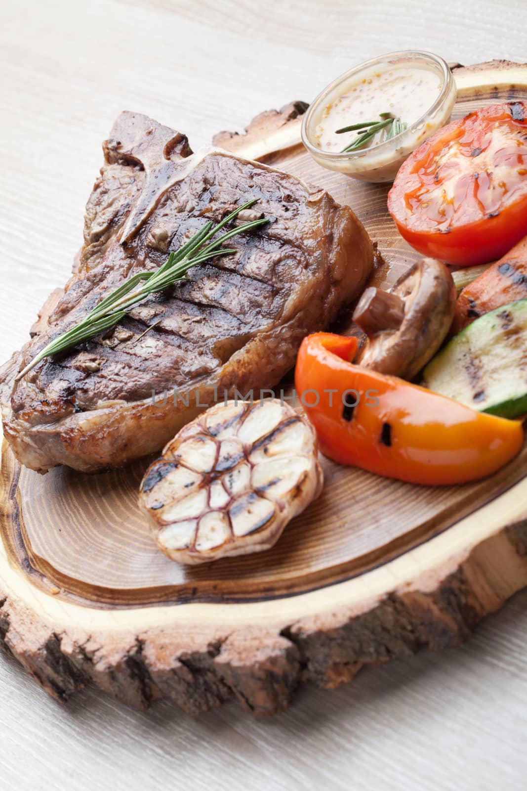 Portion of BBQ t-bone steak  served  on wooden board with  rosemary, mustard sauce  and grilled vegetables : tomato, carrot, paprika, garlic,  champignon,  zucchini