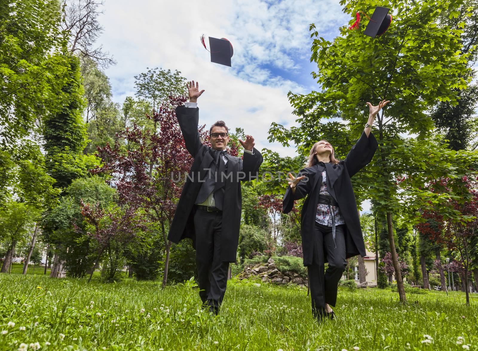 Happy couple in the graduation day running in a park and throwing  their hats up.
