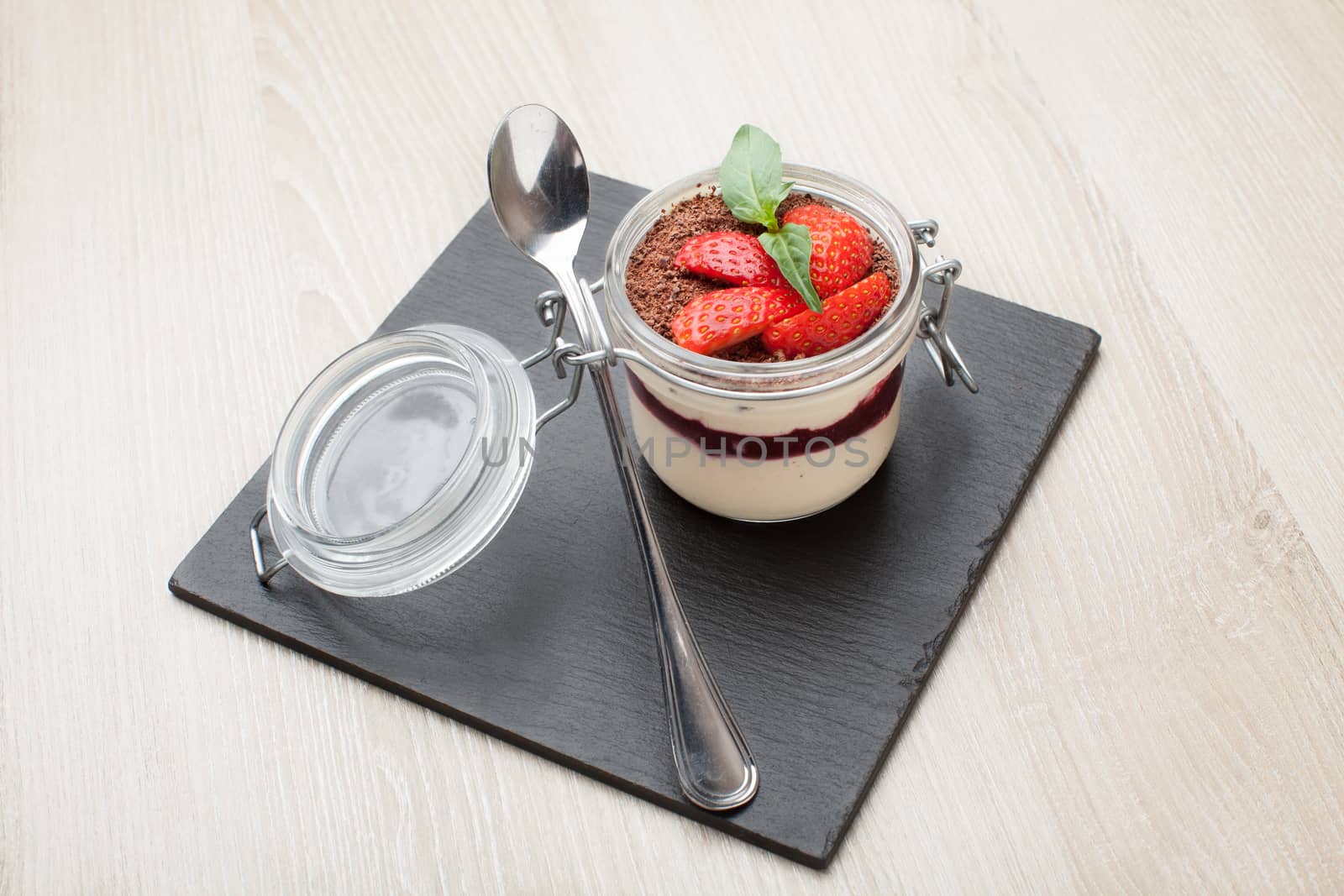 Delicious traditional italian cream dessert pannacotta with grated chocolate, strawberry, and basil served in jar with cocktail spoon on black stone background