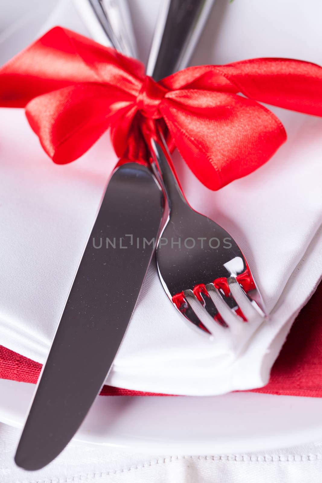 Romantic red Christmas table setting with white plates, red and white linen and silverware tied with a red ribbon and bow decorated with red Xmas baubles and evergreen natural pine foliage