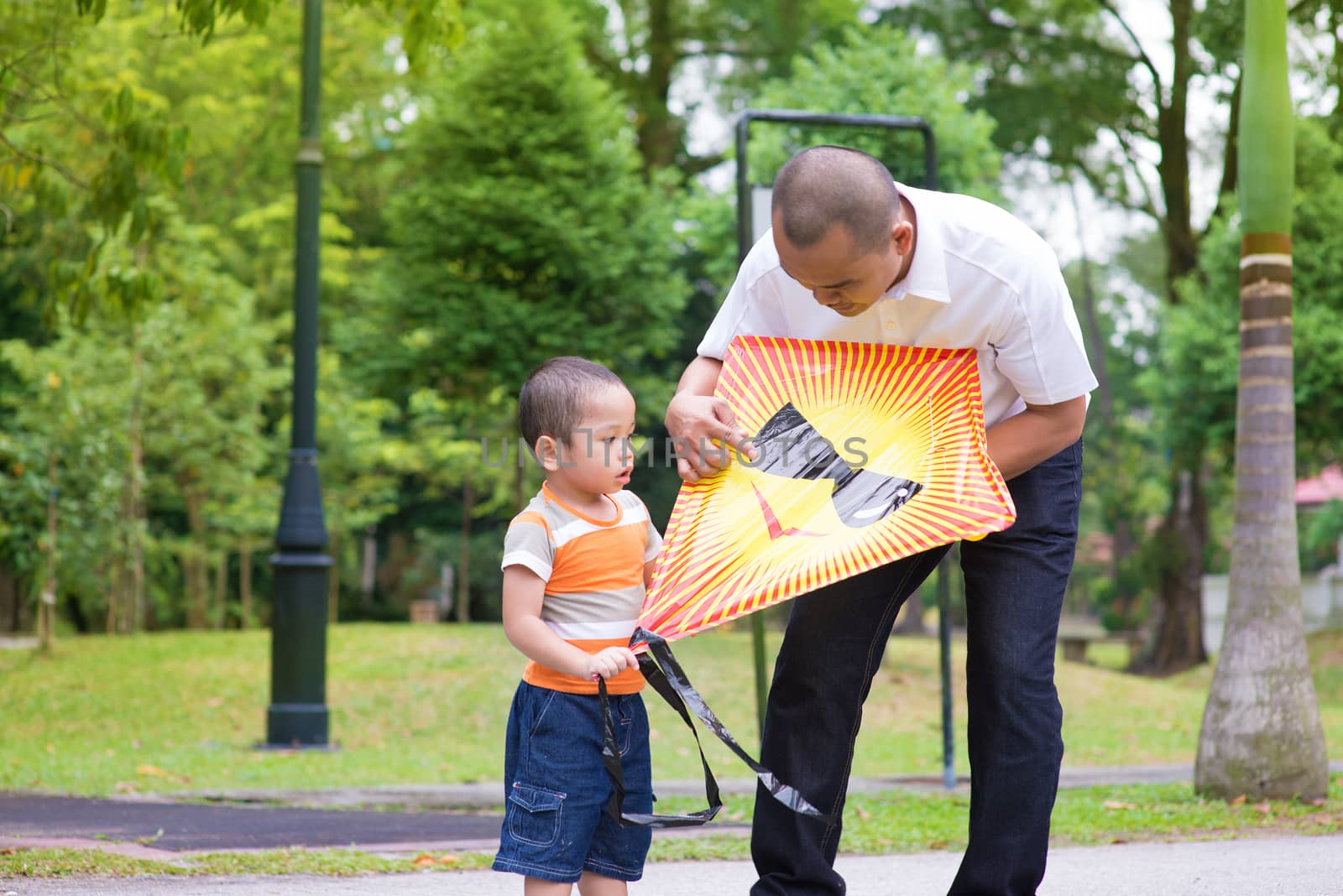 Father teaching son to fly a kite at outdoor garden park. Happy Southeast Asian family living lifestyle.
