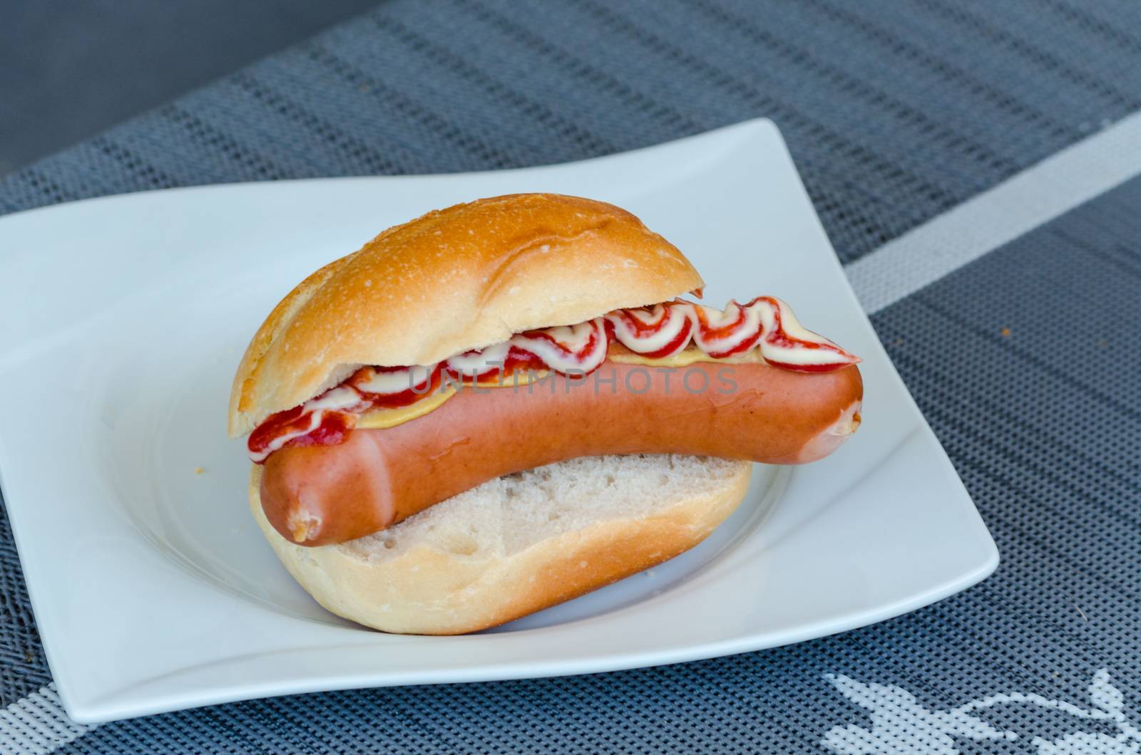 Hot dog with ketchup and mustard in bread roll on a white plate.