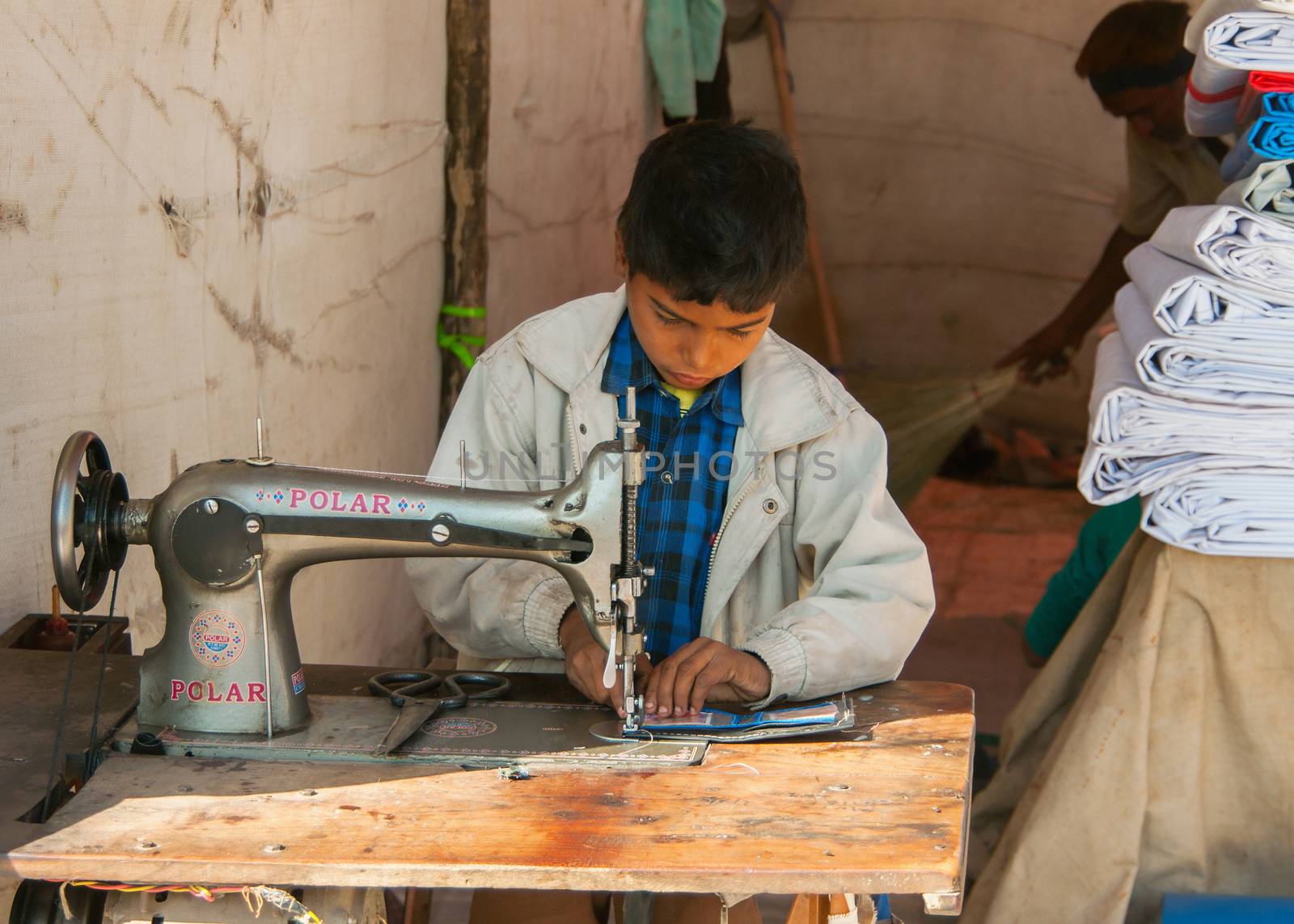 Child labor, boy sewing in booth on the market. by Claudine