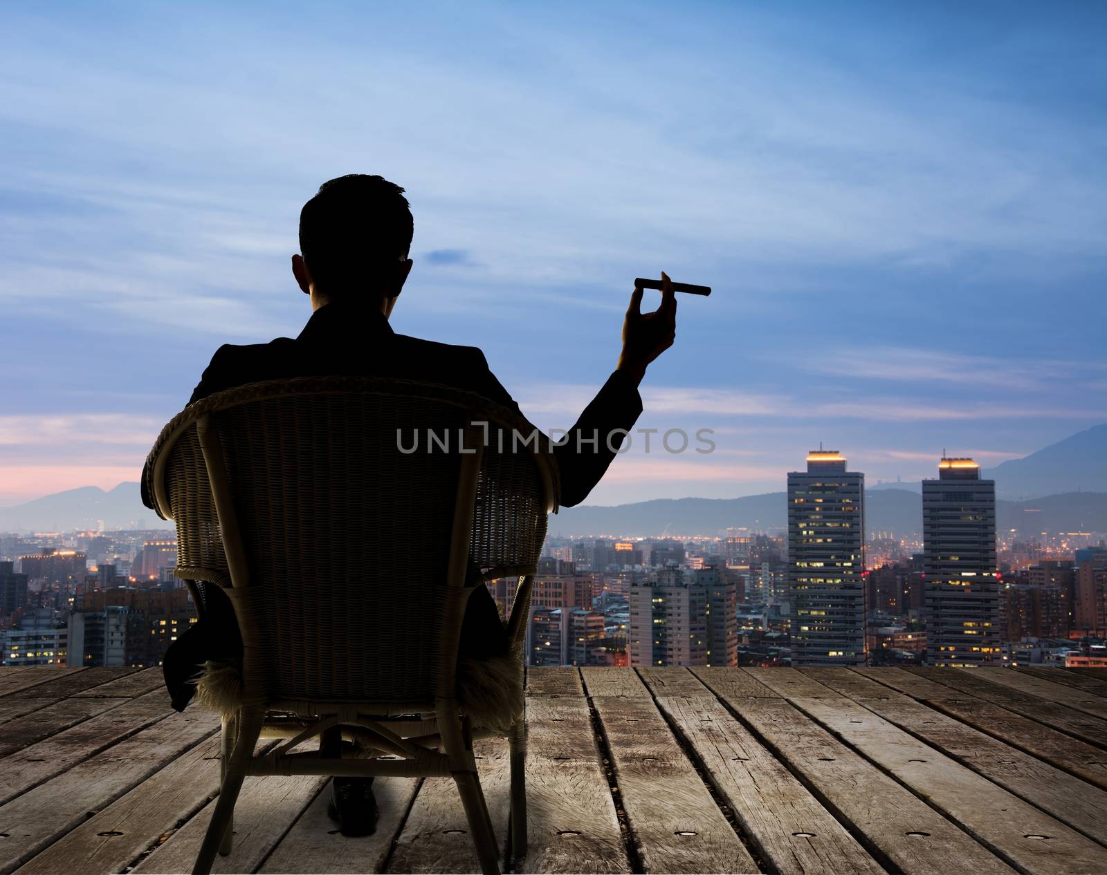 Silhouette of businessman sit on chair and hold a cigar and looking at the city in night.