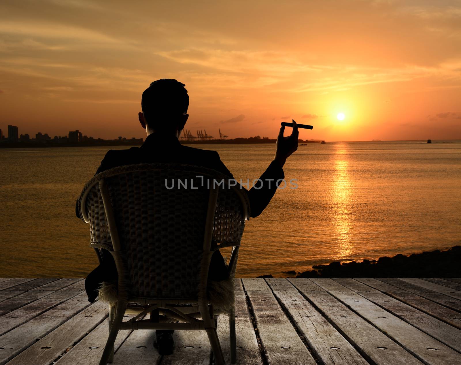 Silhouette of businessman sit on chair and hold a cigar and looking at the city in night.