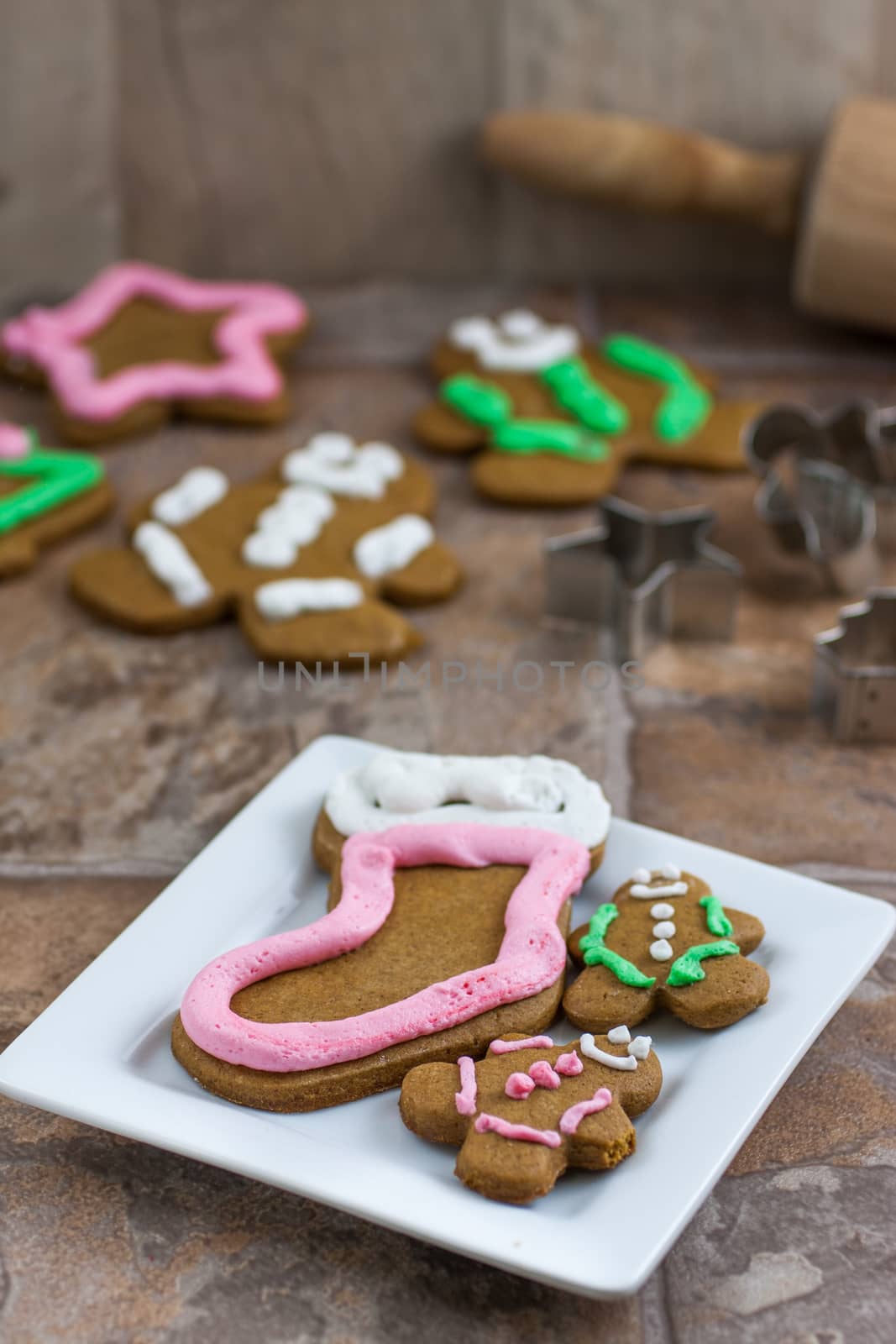 Several gingerbread Christmas cookies on a white plate with cookie cutters, a rolling pin, and several cookies in the background.