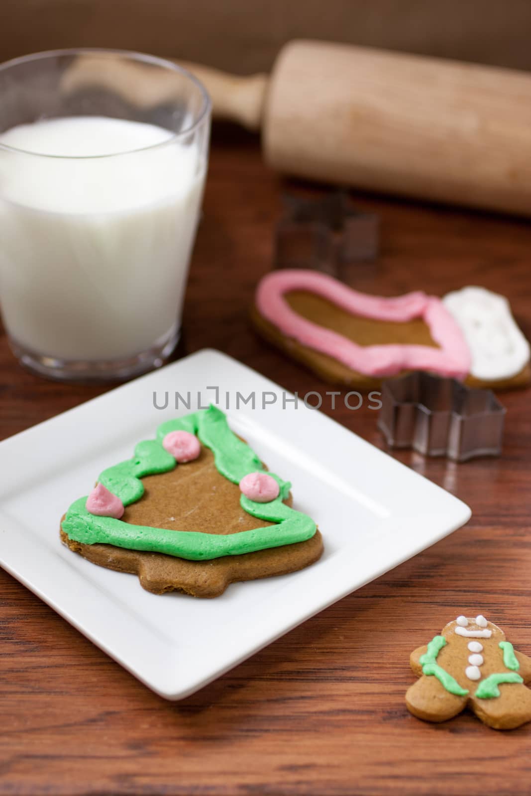 Several gingerbread Christmas cookies on a white plate with cookie cutters, a rolling pin, and several cookies in the background.