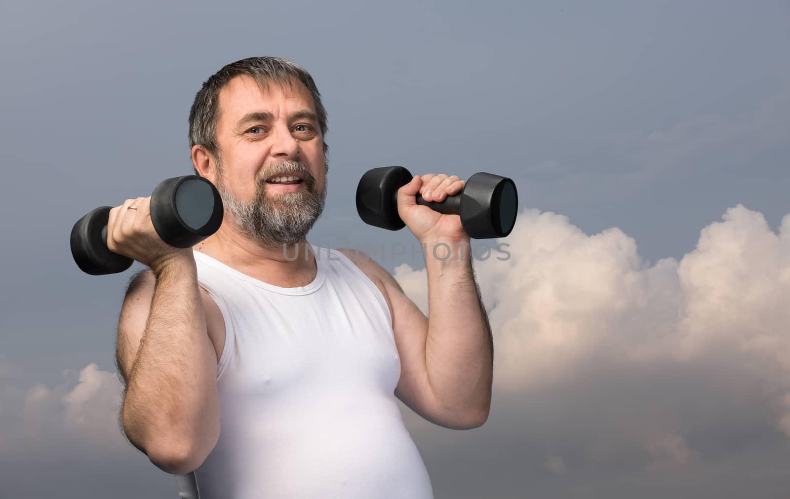 Elderly man exercising with dumbbells against cloudy sky background