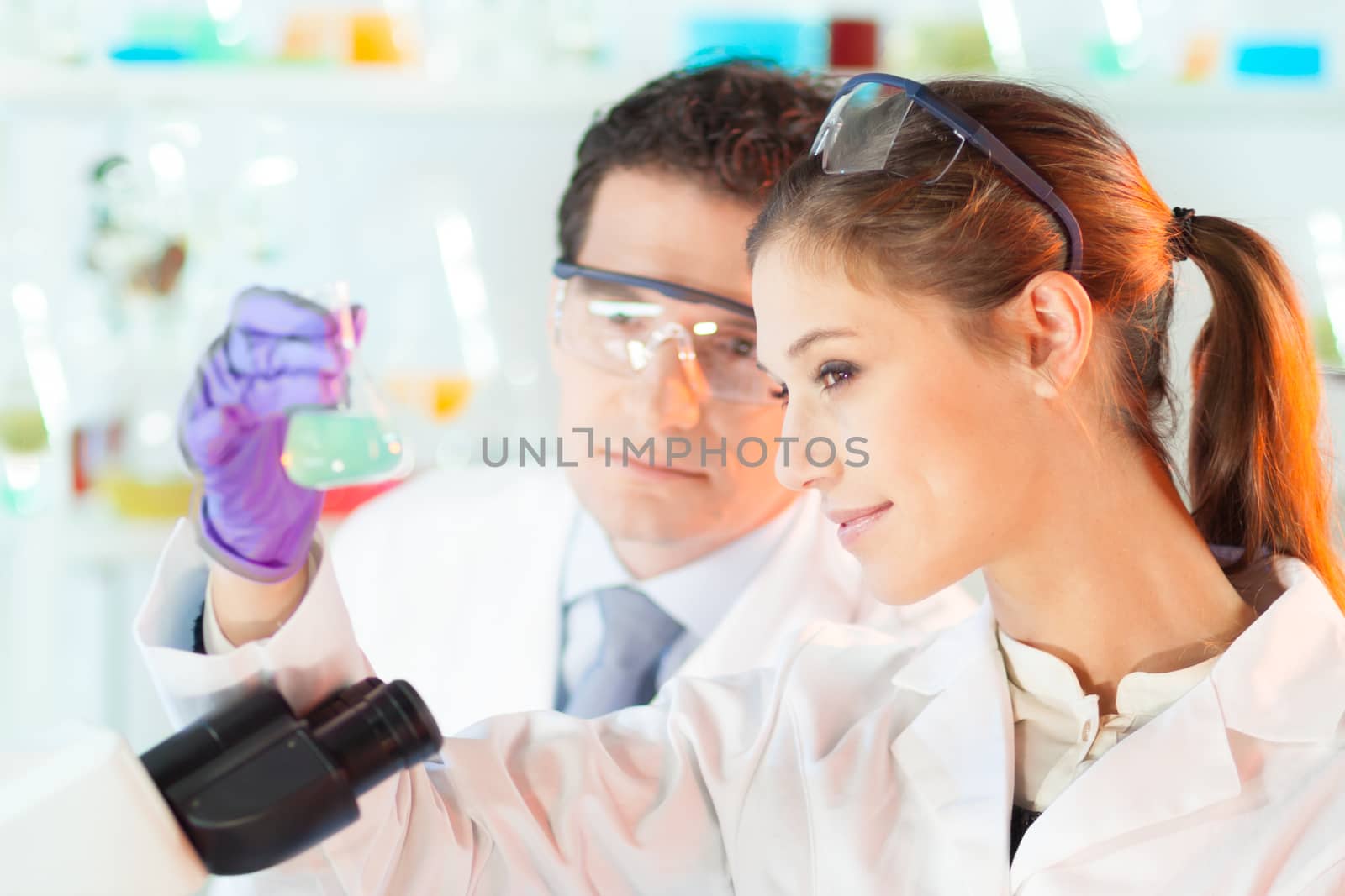 Chemical laboratory scene: attractive young student and her post doctoral supervisor scientist observing the green indikator solution color shift in glass flask.