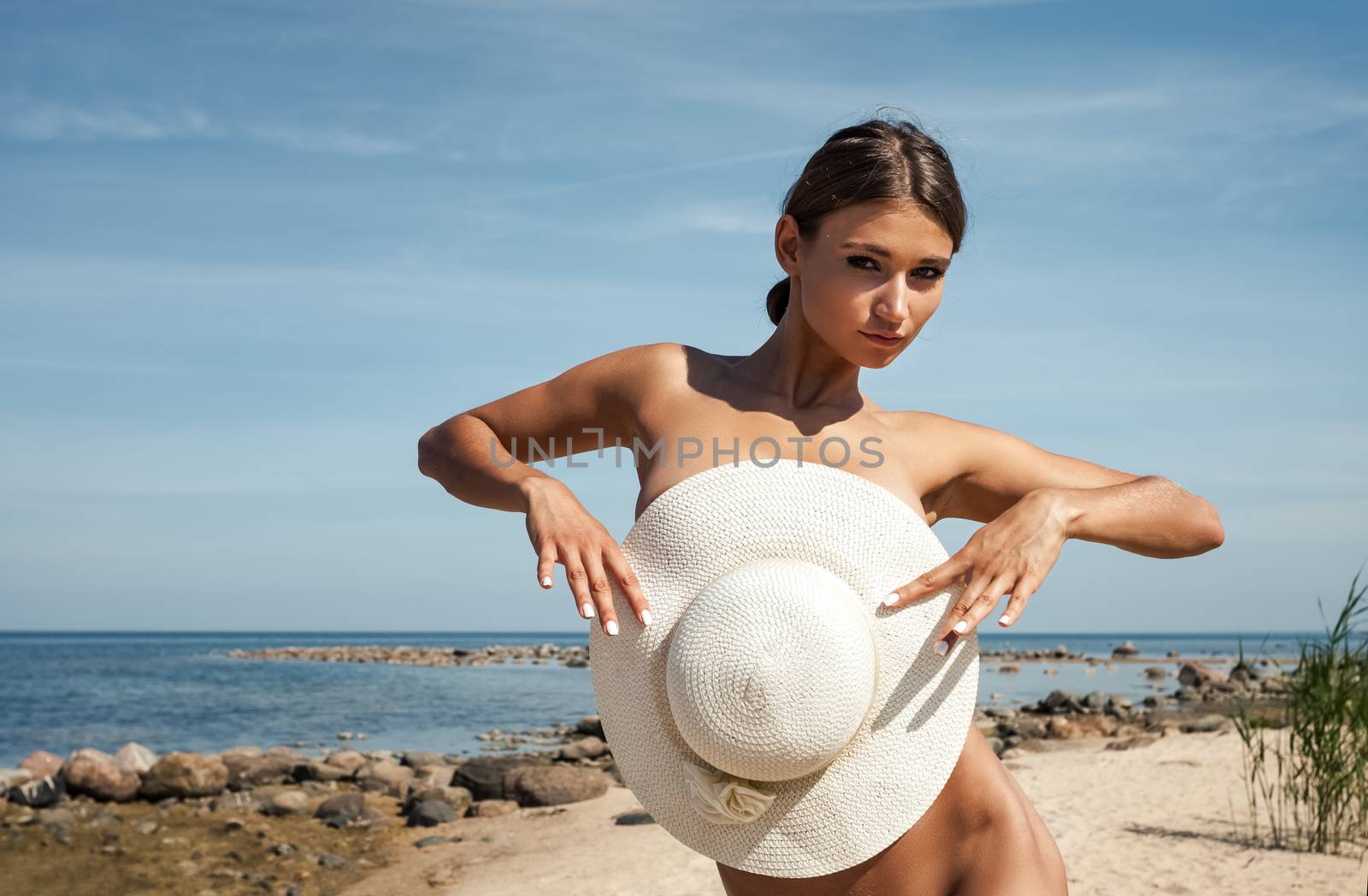 Young elegant nude woman with a white hat posing on the beach
