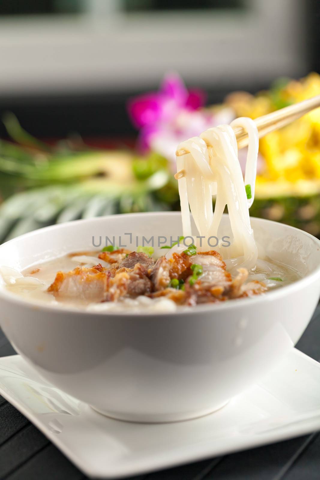 Closeup of a person eating Thai style crispy pork rice noodle soup from a bowl with chopsticks. Pineapple fried rice in the background.