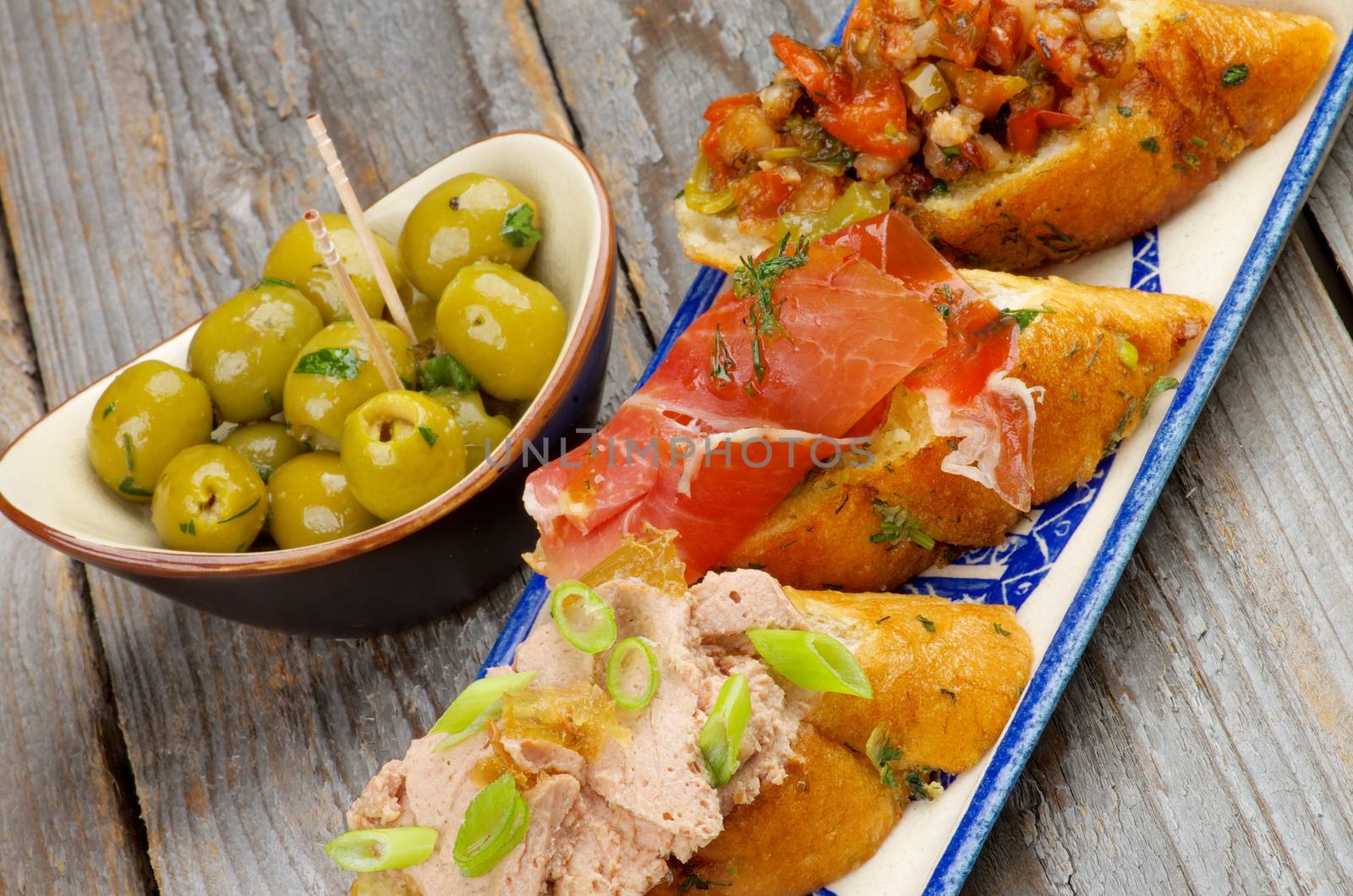 Tapas Bruschetta with Pate and Spring Onion, Bacon, Roasted Vegetables and Cured Ham with Green Olives closeup on Rustic Wooden background