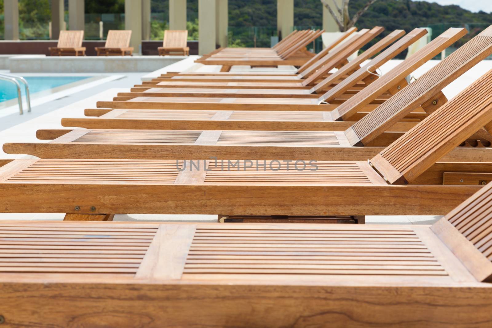 modern wooden sunbeds by turquoise swimming pool. 