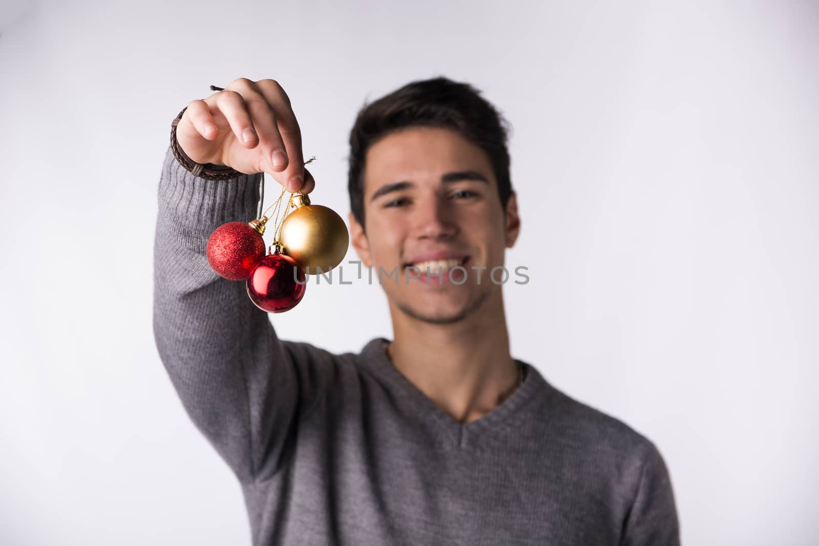 Handsome young man smiling and holding Christmas tree balls in front of his face, looking at camera on white background