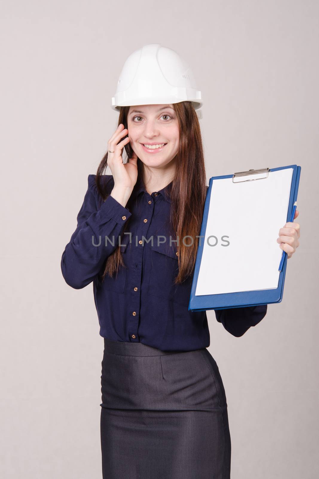 Young woman builder in helmet standing with a folder in hand and talking on phone