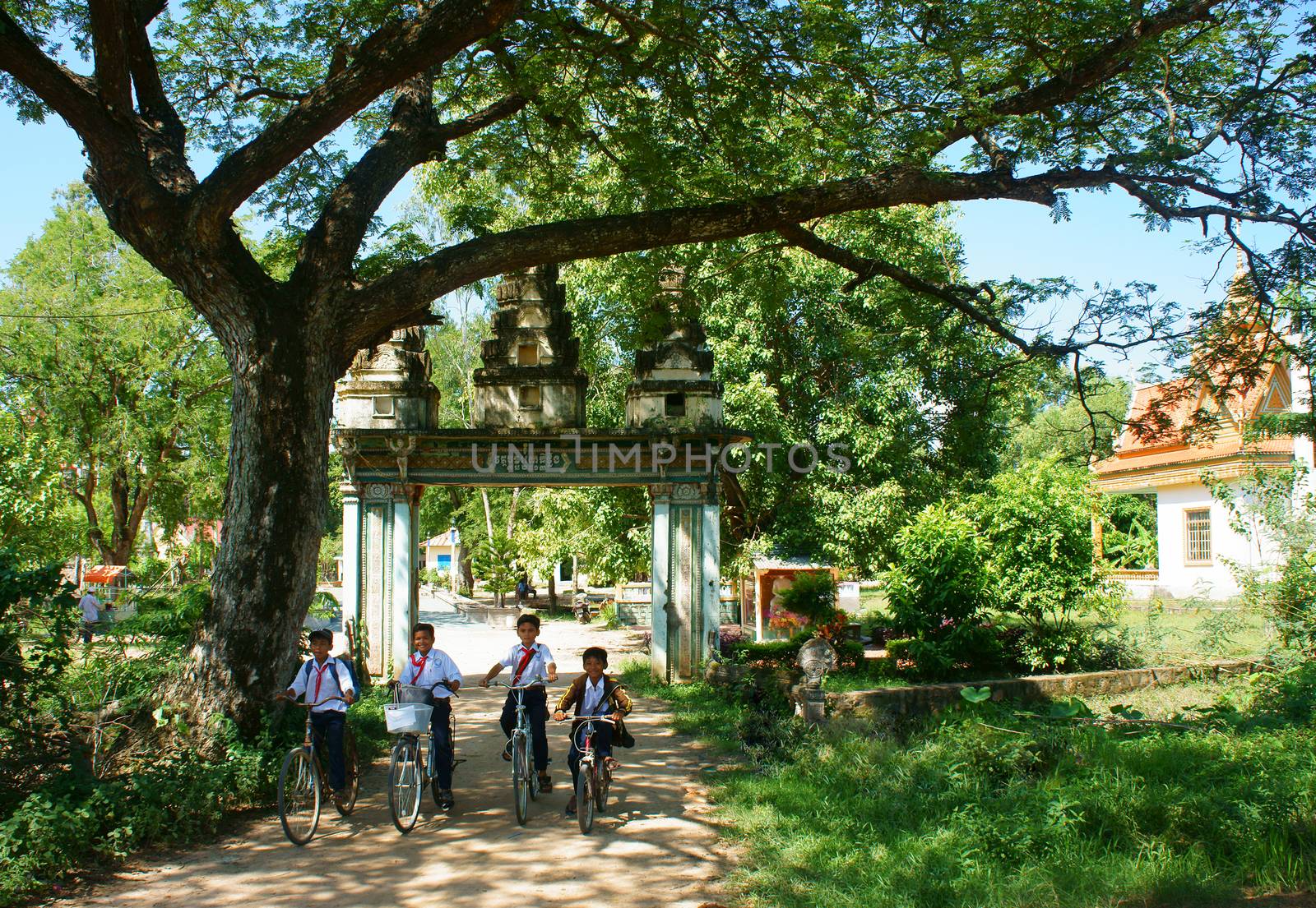 AN GIANG, VIET NAM- SEPT 20: Group of Asian primary pupil riding bike to Khmer village gate, ancient gate with large green tree, big brach make shade, kid happy with friendship, Vietnam, Sept 20, 2014