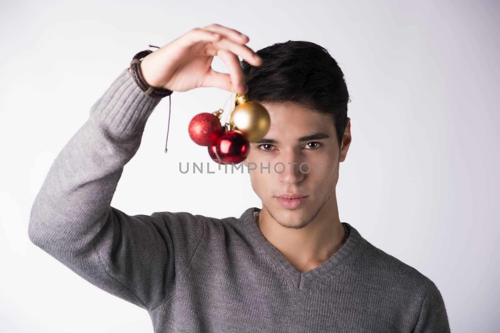 Handsome young man holding Christmas tree balls in front of his face, looking at camera on white background