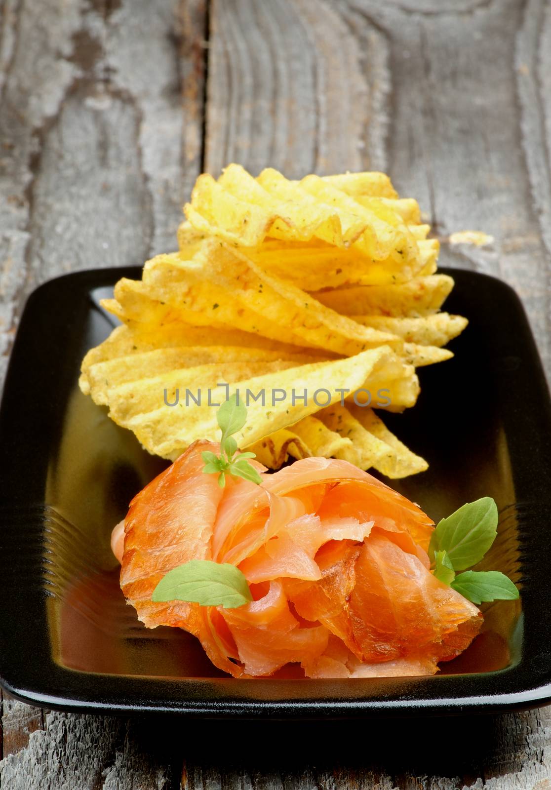 Delicious Smoked Salmon with Rifled Potato Chips closeup on Black Plate on Rustic Wooden background
