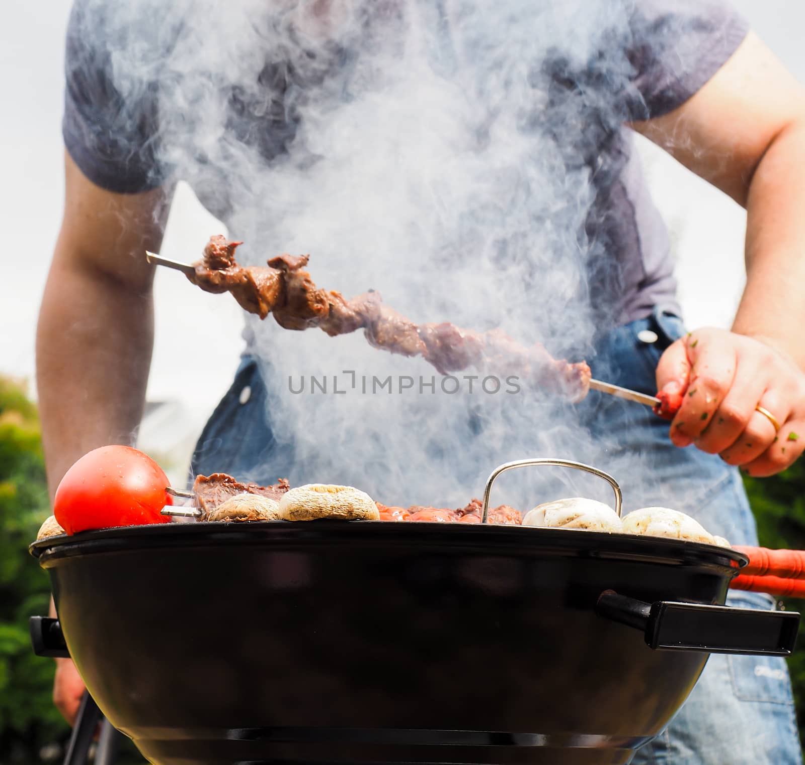 Chef covered in smoke grilling skewers of meat and vegetables over charcoal barbecue