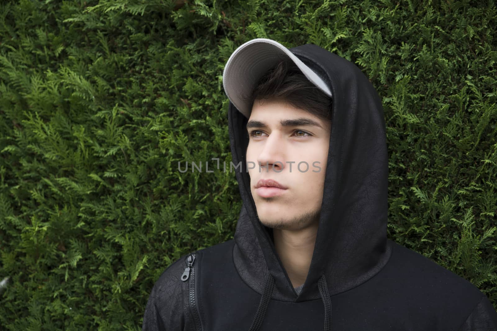 Attractive young man with hoodie and baseball cap against green bush "wall", sad or thinking