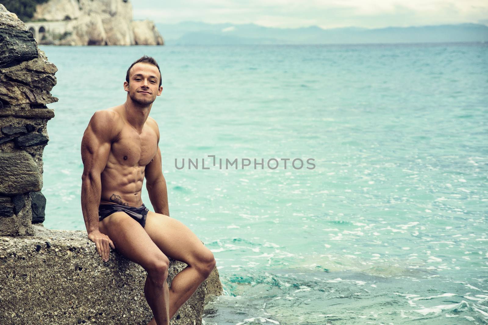 Handsome muscular young man on the beach sitting on rocks, smiling at camera