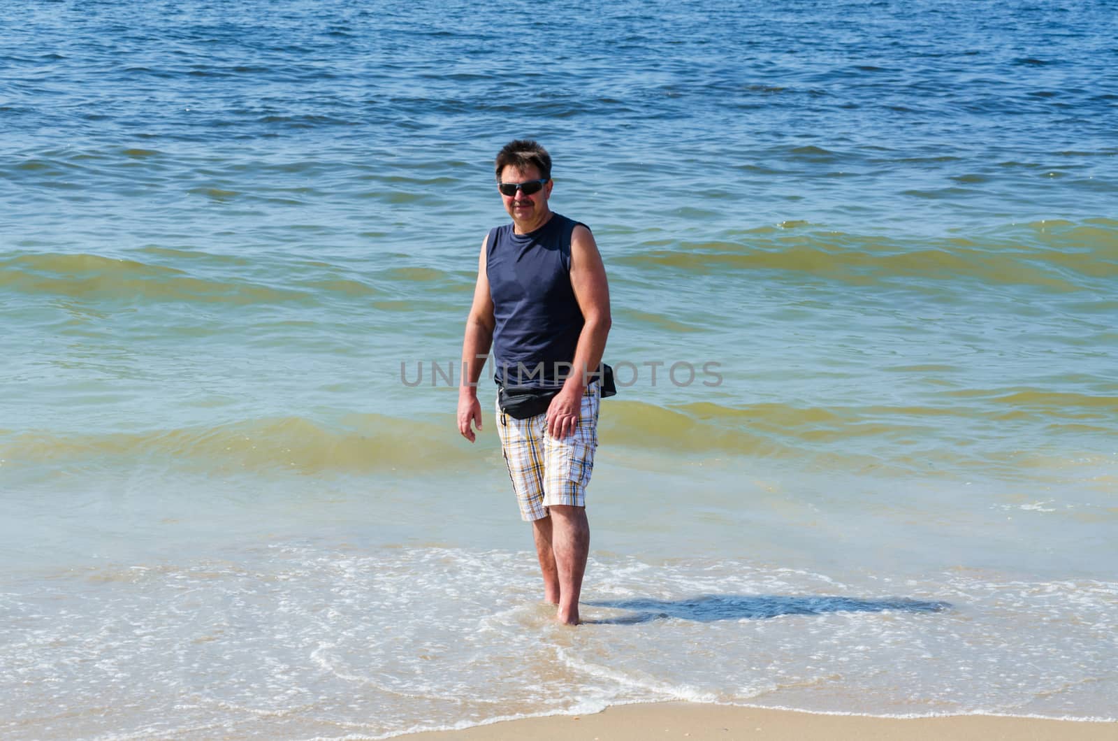 Man in shorts, T-shirt and glasses at the beach with your feet in the water.