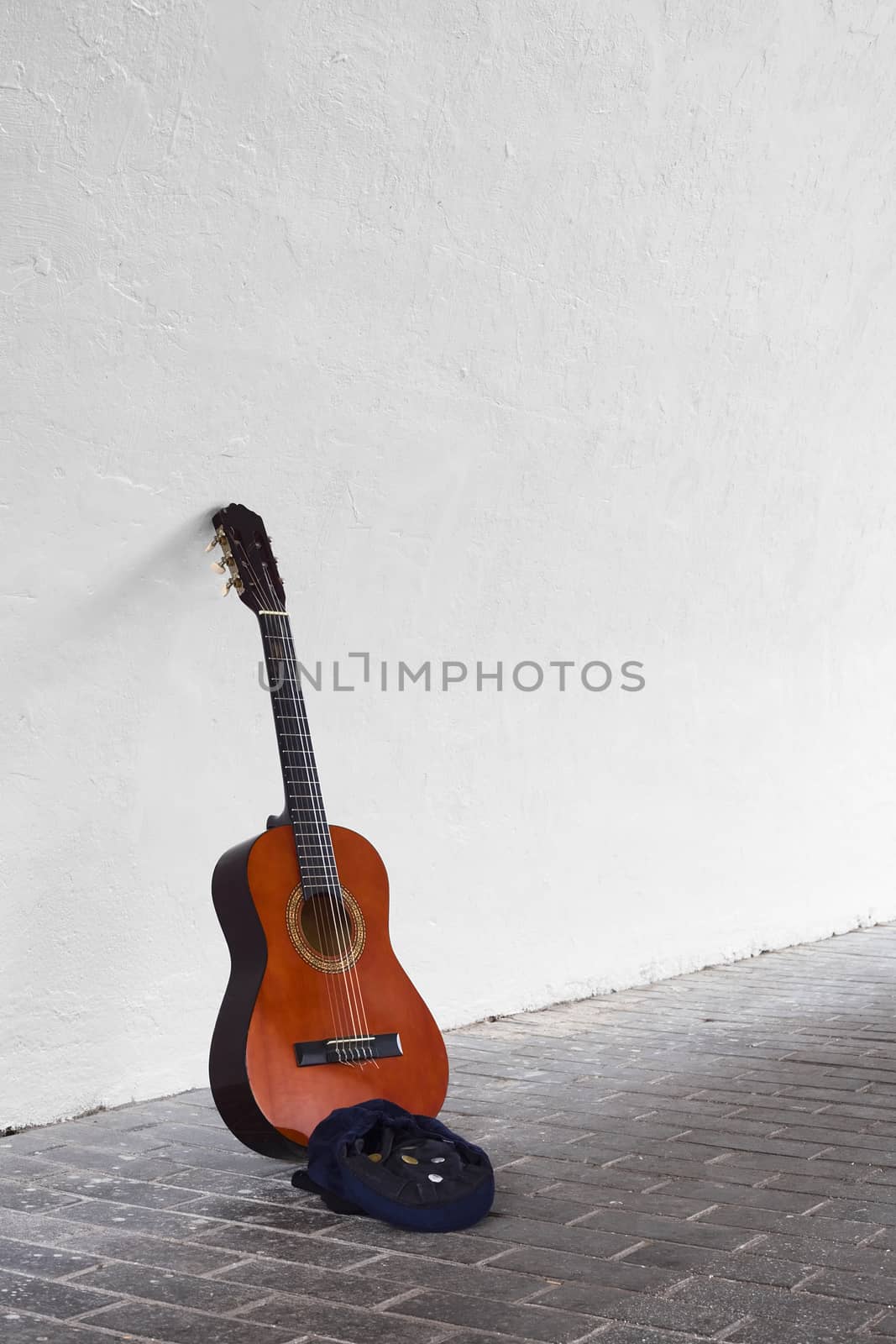Guitar leaning against white wall with blue hat containing some coins in front 