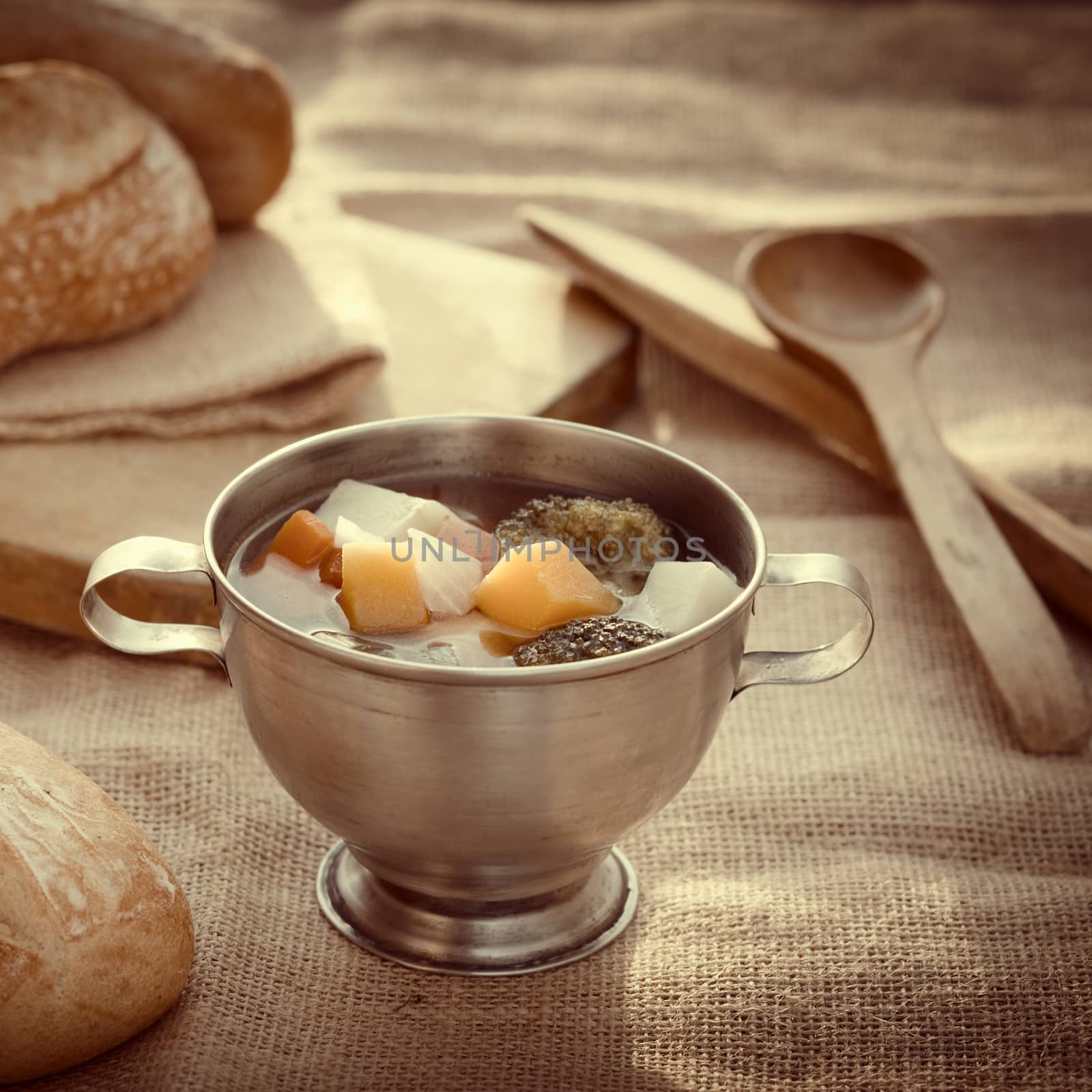 Fresh vegetarian soup made of broccoli, squash, onion, carrot, potato and tomato served in a metal cup on jute cloth and accompanied by ciabatta buns, low contrast image (Selective Focus, Focus on the middle of the soup)