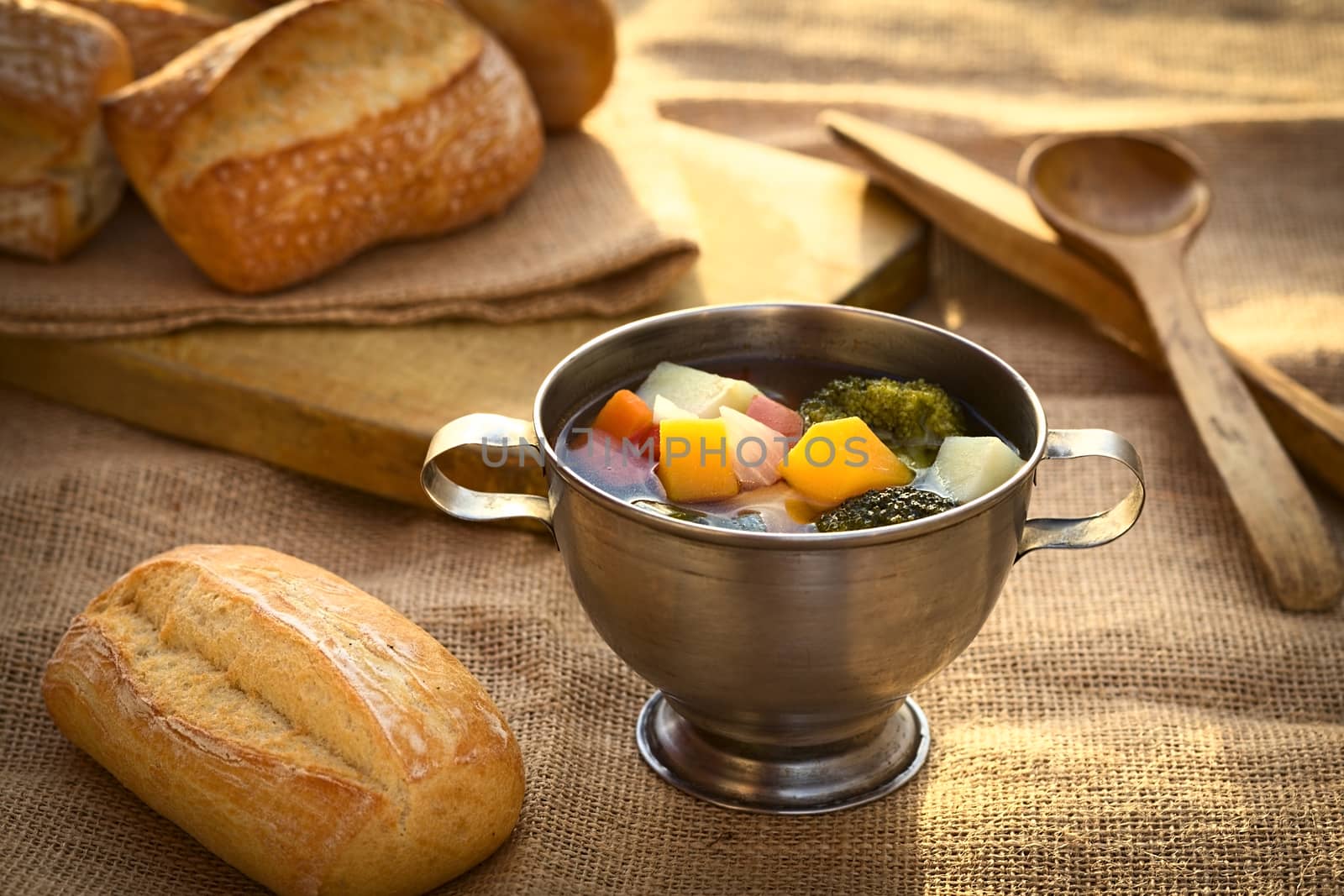 Fresh vegetarian soup made of broccoli, squash, onion, carrot, potato and tomato served in a metal cup on jute cloth and accompanied by ciabatta buns, photographed with natural light (Selective Focus, Focus onto the first half of the soup)