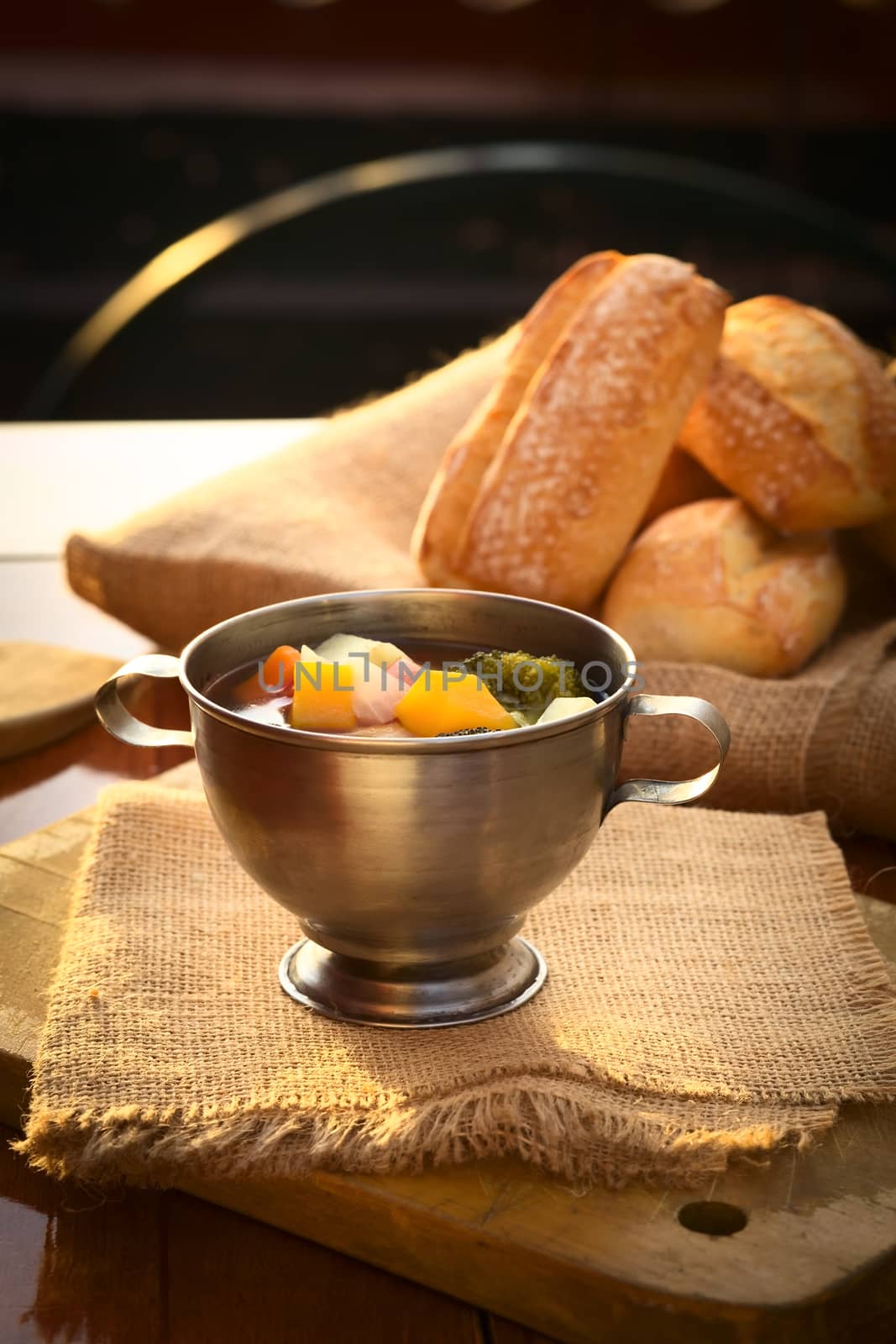 Fresh vegetarian soup made of broccoli, squash, onion, carrot, potato and tomato served in a metal cup on jute cloth and wooden board with ciabatta buns in the back, photographed with natural light (Selective Focus, Focus on the squash pieces in the middle of the soup)