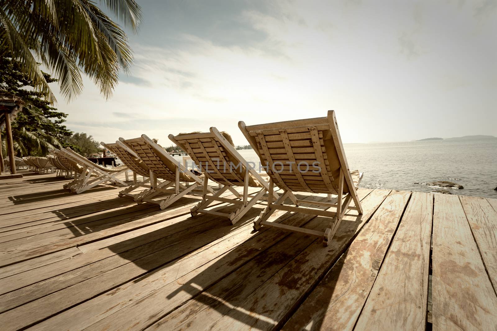 Vacation Concept. Palms and relaxing sunbeds on wooden desk near the sea under blue sky. Sepia toned
