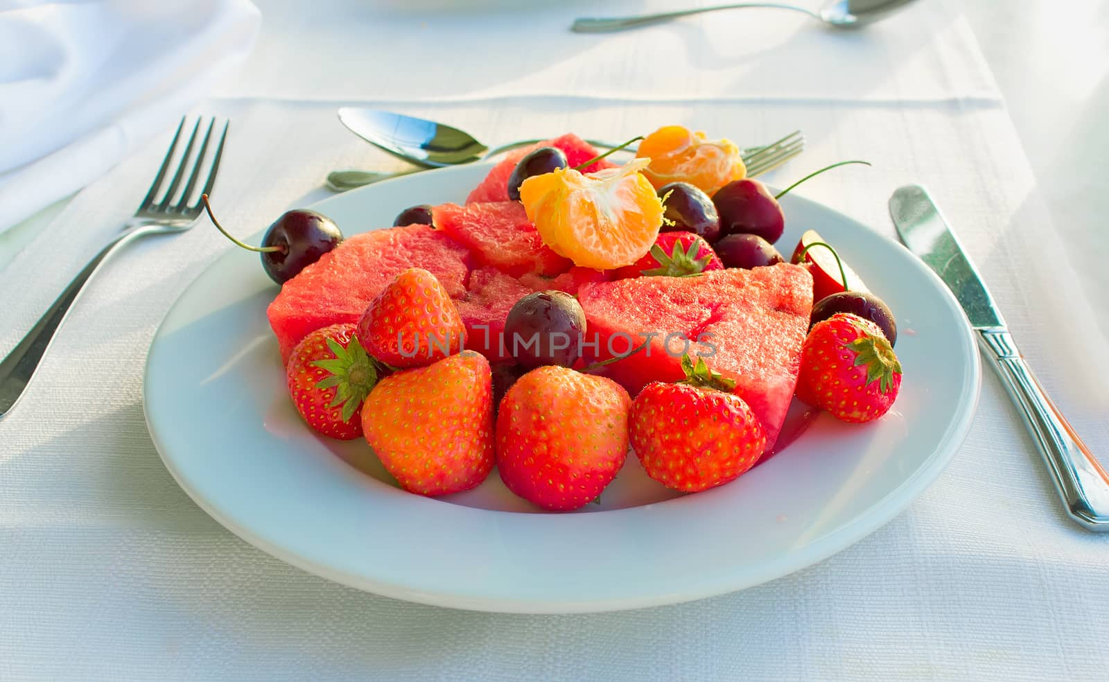 Strawberries, cherries, slices of watermelon, slices of Mandarin are located on a plate on a white tablecloth table.