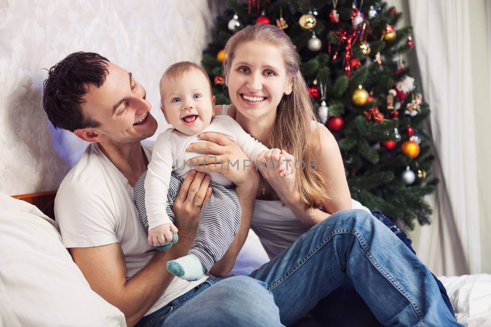 Young family with a baby having fun on bed at home with Christmas tree in the background