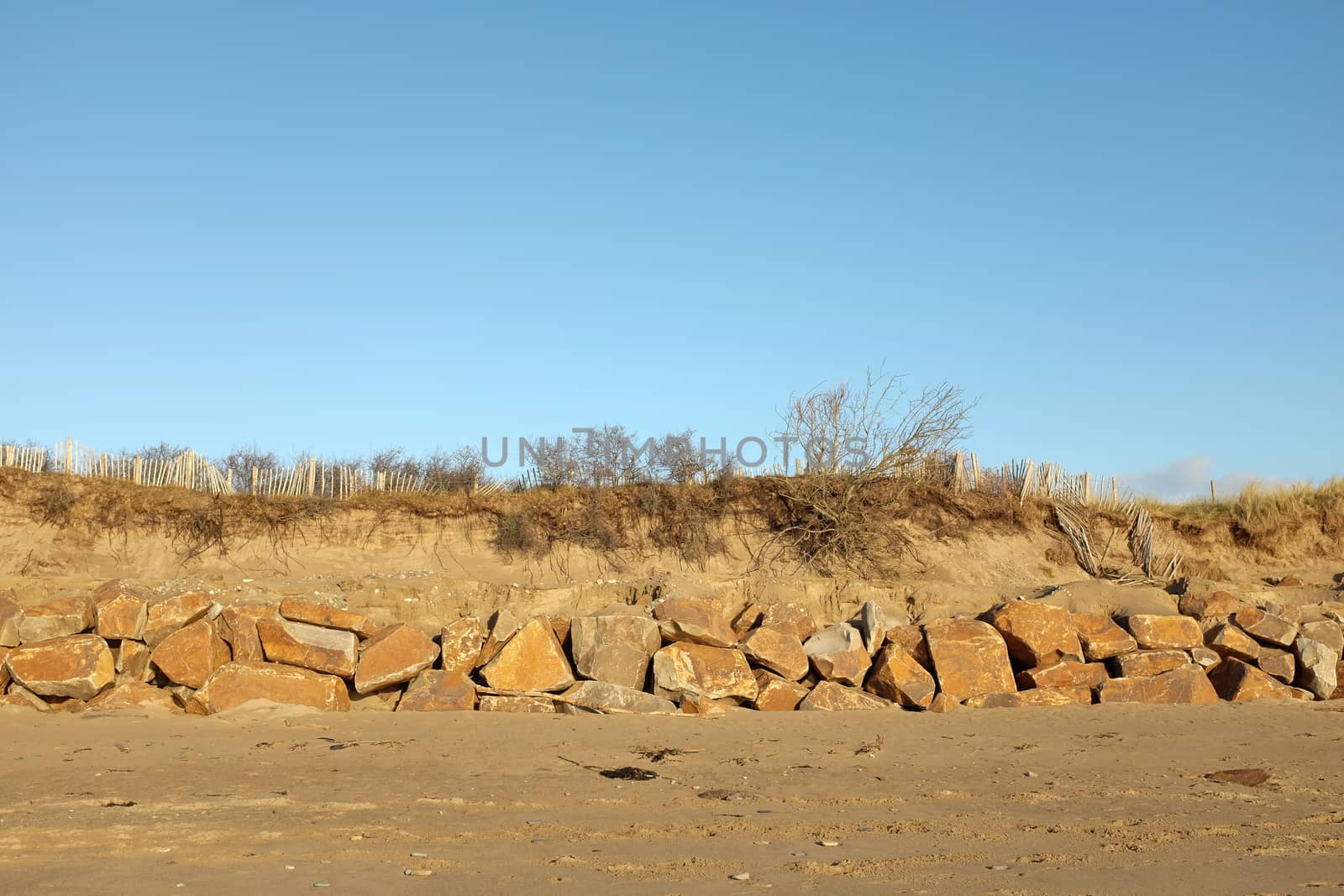 A sandy beach lead to a row of large rocks with an eroded sand bank in the rear and blue sky in the distance.