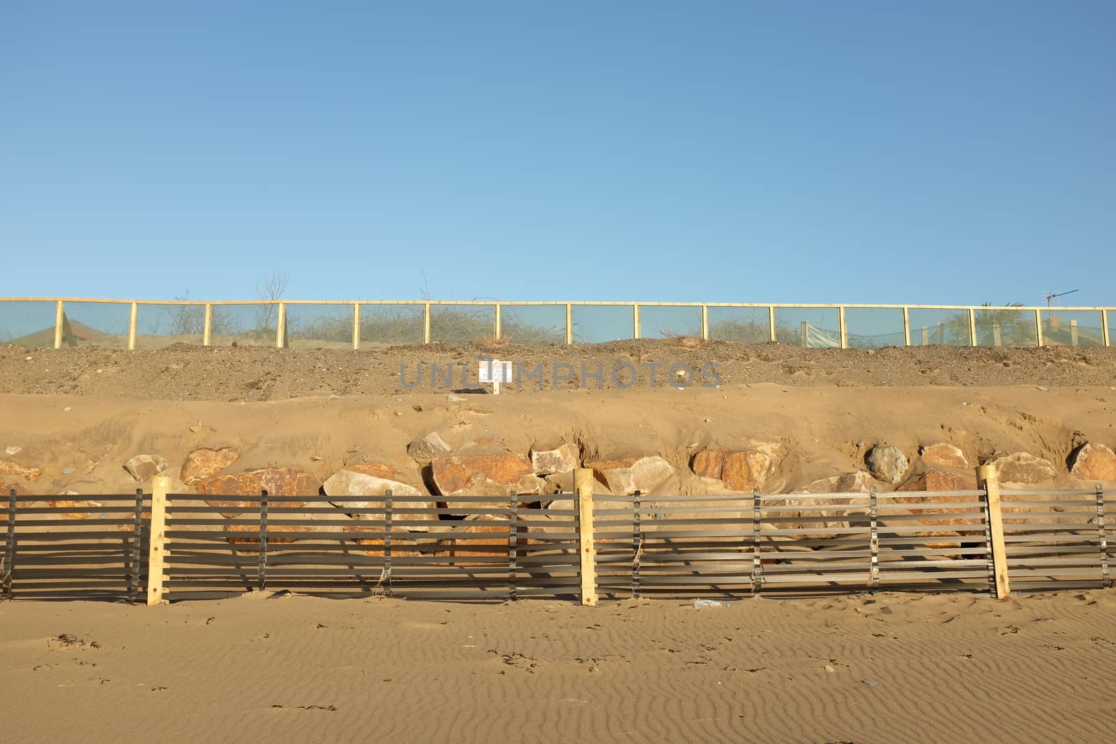 An area of constructed sand dune with a sand drift fence on wooden posts in front of a row of large rocks banked up with sand, a danger sign and a protective fence on top.