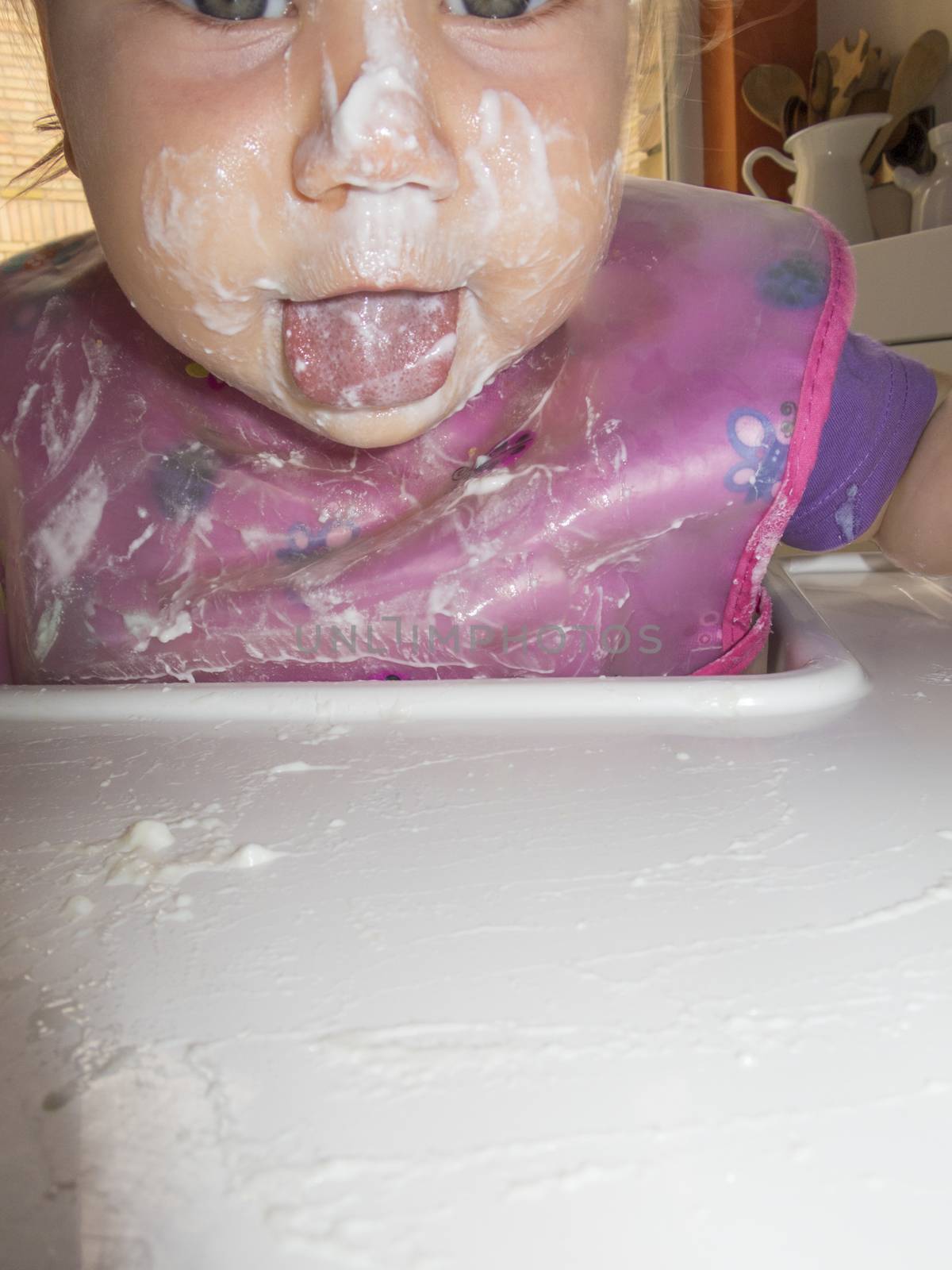 caucasian baby 1 year eating natural yogurt with her hand in high-chair kitchen