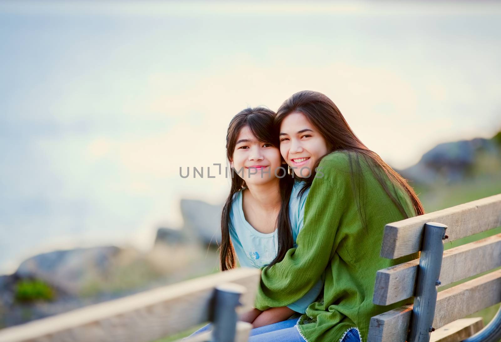 Two happy biracial young girls sitting together on bench next to lake