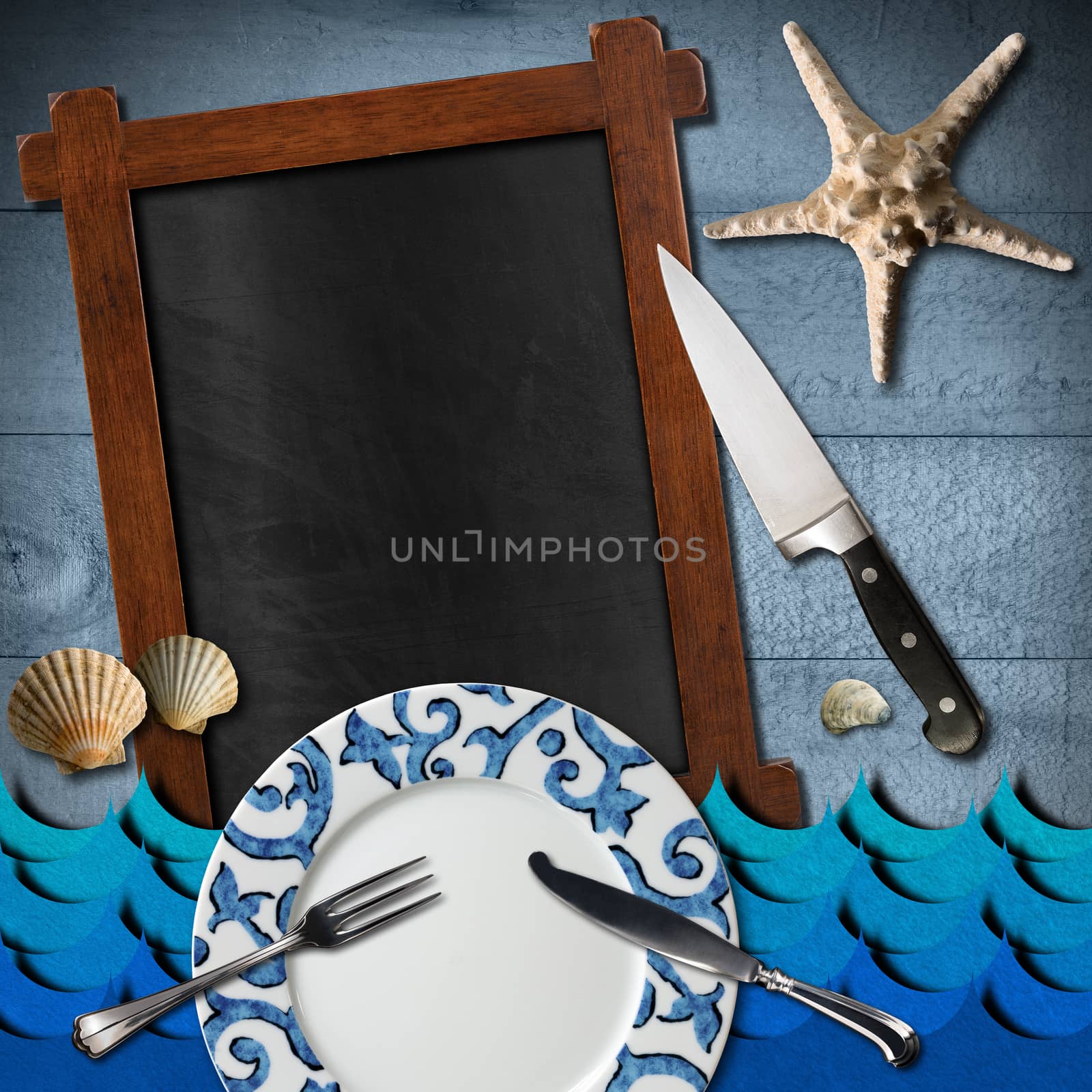 Blackboard with wooden frame, stylized sea waves, empty plate and cutlery, kitchen knife, seashells and starfish on blue wooden background. Template for recipes or seafood menu