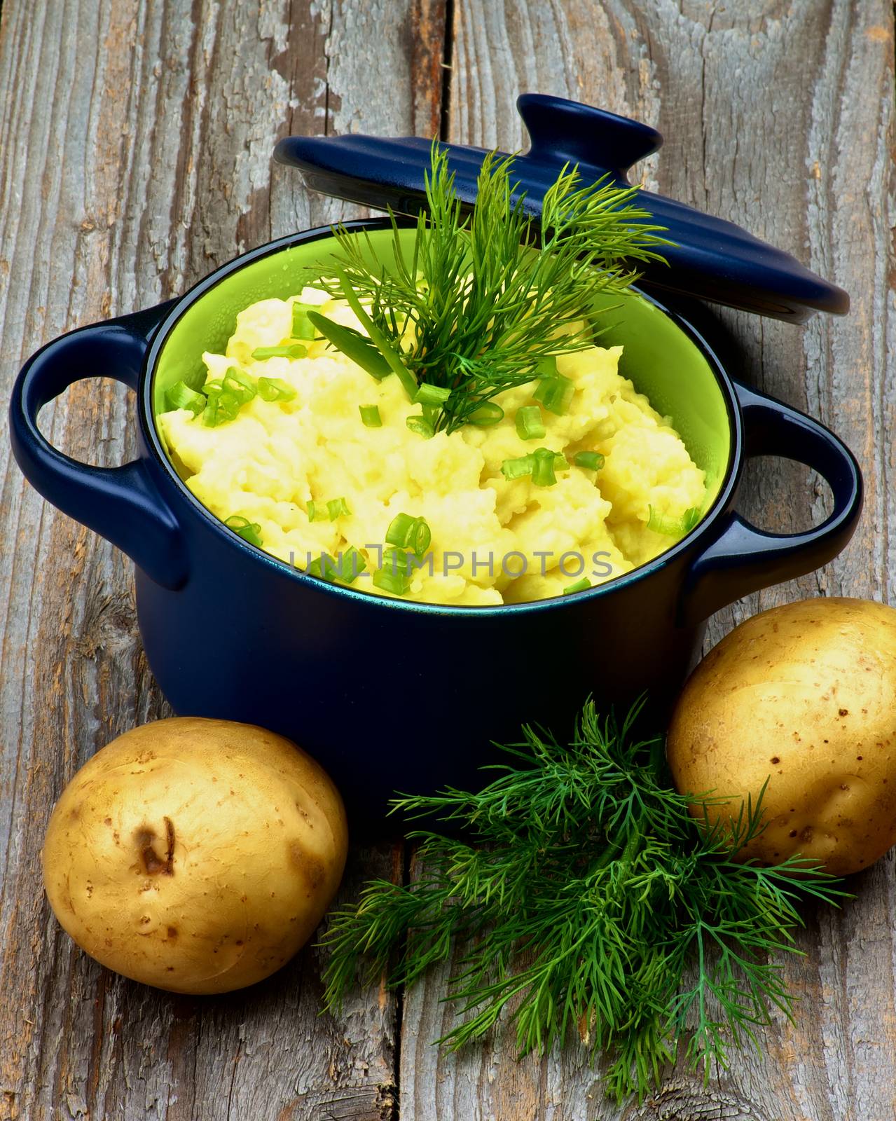 Creamy Homemade Mashed Potato with Dill and Spring Onion in Dark Blue Casserole with Lid and Raw Potatoes on Rustic Wooden background