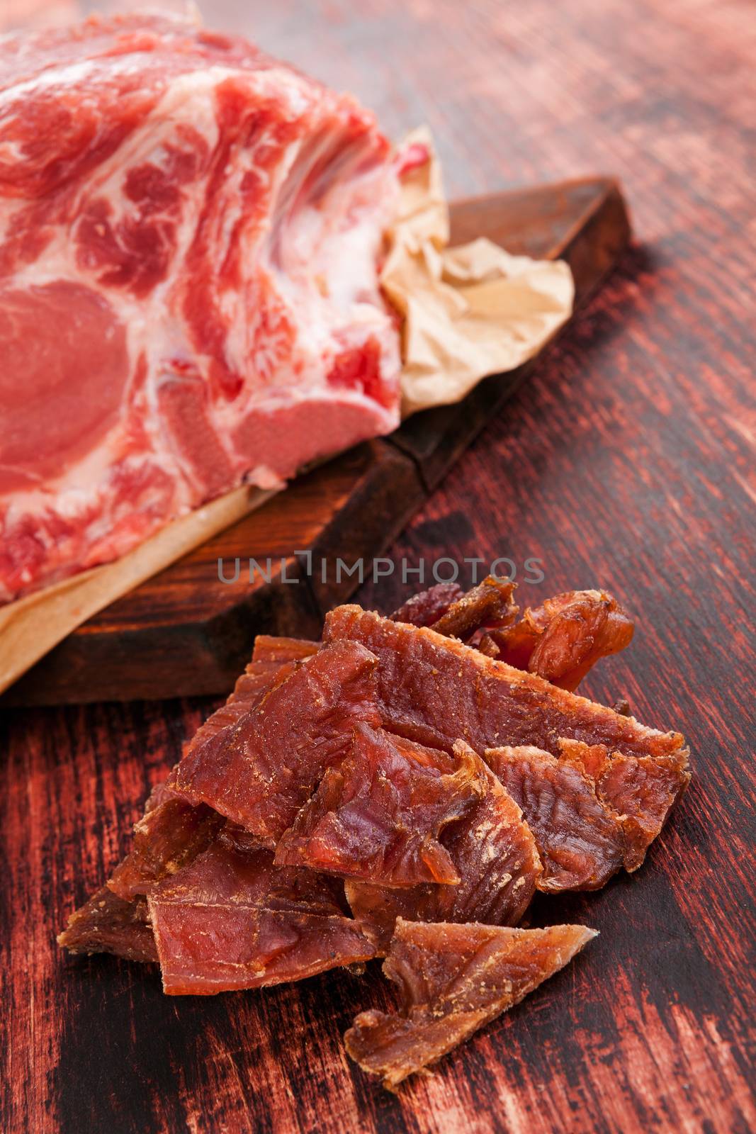 Dry meat, beef jerky. Beef jerky pieces and big fresh meat piece on brown wooden background. Meat eating, rustic country style.