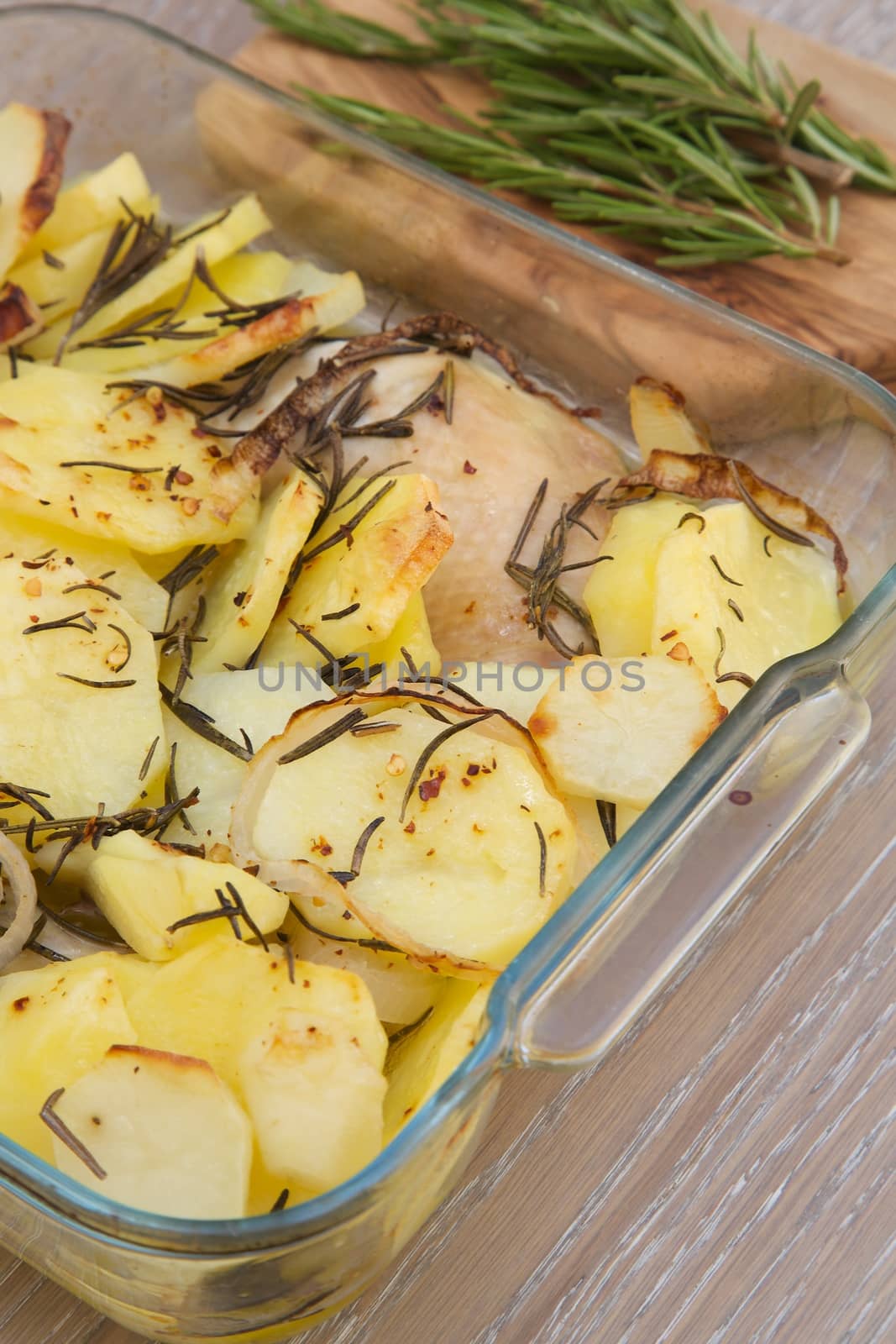 Baked potatoes with chicken and rosemary in the glass mould. Fresh rosemary on the wooden cutting board in the background