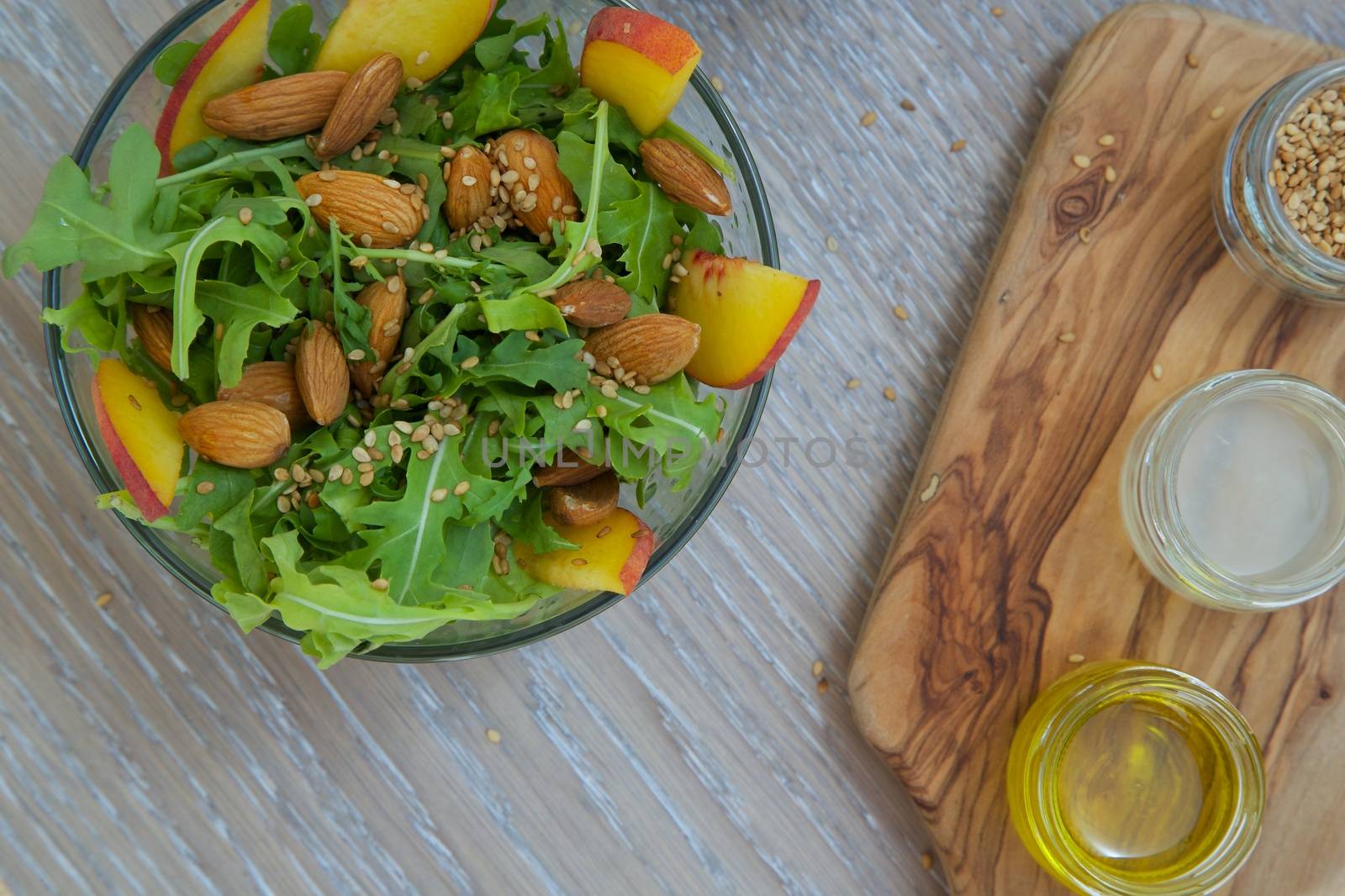 Vitamin salad- rucola with almond, peach and sesame seeds in glass dish. Olive oil, lemon juice and sesame seeds on the wooden cutting board in the background. Close up