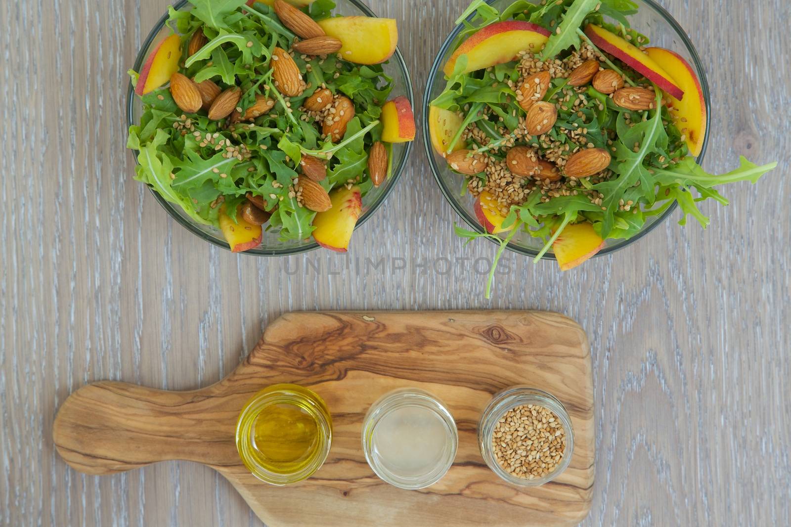 Vitamin salad- rucola with almond, peach and sesame seeds in glass dishes. Olive oil, lemon juice and sesame seeds on the wooden cutting board. Background. Top view