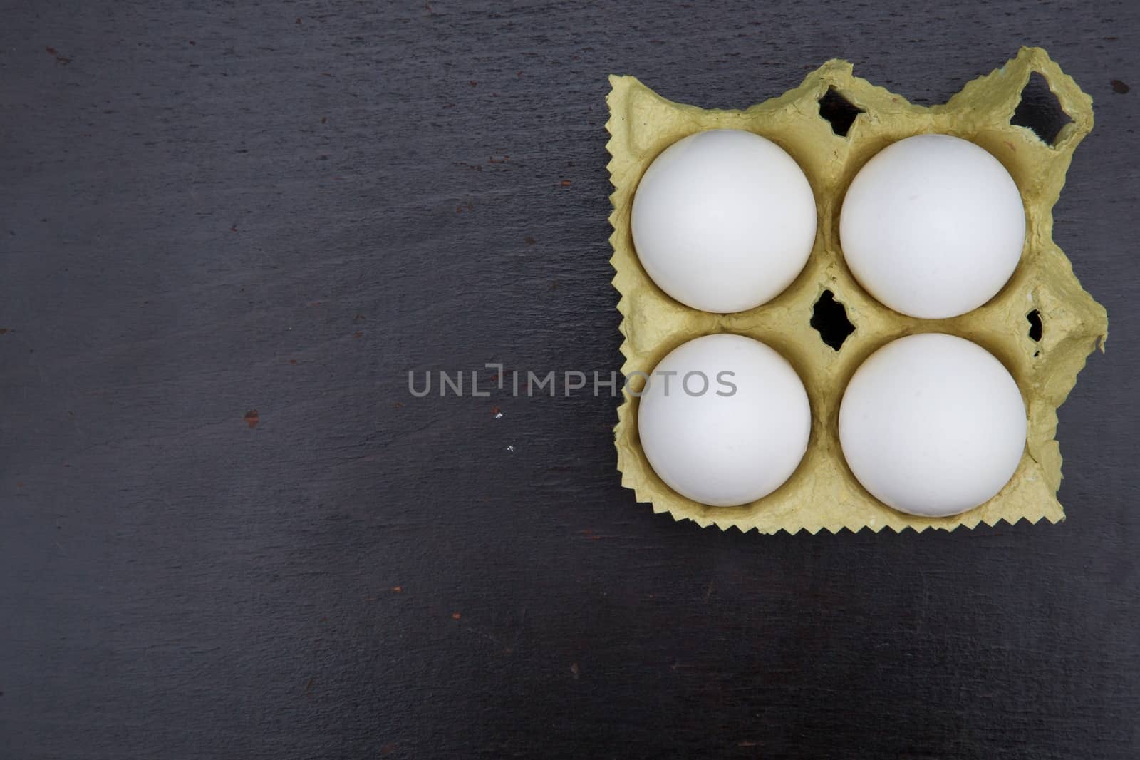 Four white chicken eggs in the carton box on the black wooden surface. Free space for a text.