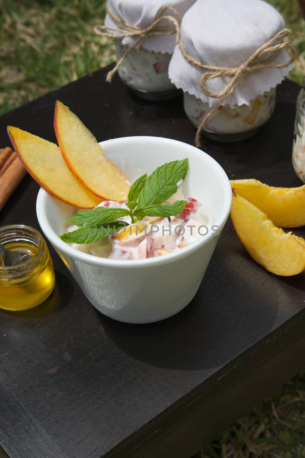 Homemade yogurt with pieces of fresh peach and cinnamon in a white dish on a black wooden surface. Pieces of peach, honey in the glass, cinnamon sticks and peach yogurt glasses in the background